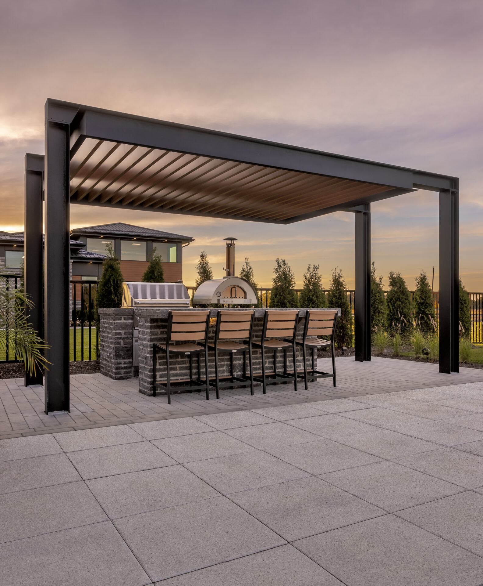 Modern outdoor seating area under a pergola, featuring a pizza oven and bar counter, with a sunset view and landscaped greenery.