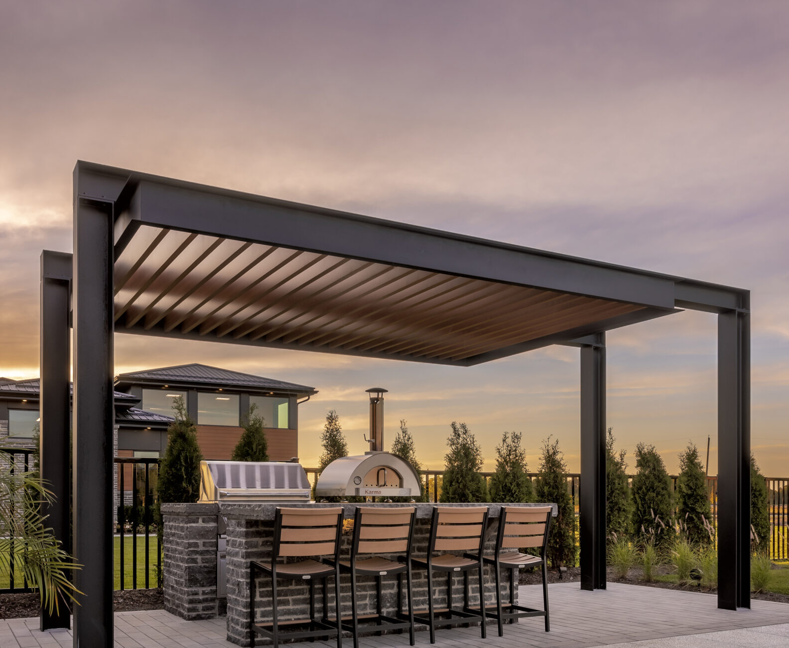 Modern outdoor patio with pergola, grill, and pizza oven. Surrounded by neatly trimmed bushes, chairs, and a house in the background. Sunset lighting.
