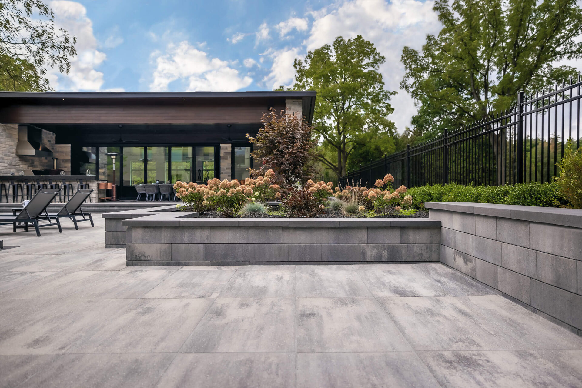 Modern patio with lounge chairs, lush landscaping, and sleek building. Open space ideal for relaxation, bordered by trees and a black metal fence.