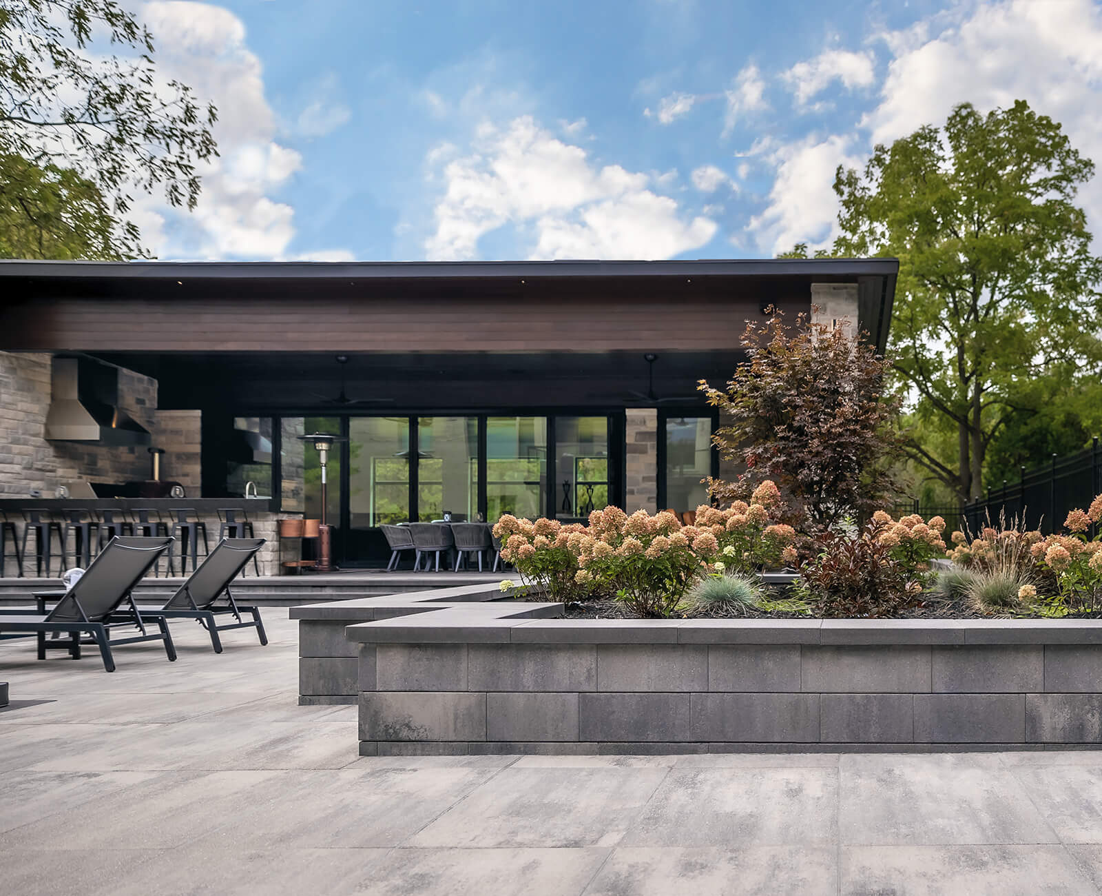 Modern patio with lounge chairs, lush landscaping, and sleek building. Open space ideal for relaxation, bordered by trees and a black metal fence.