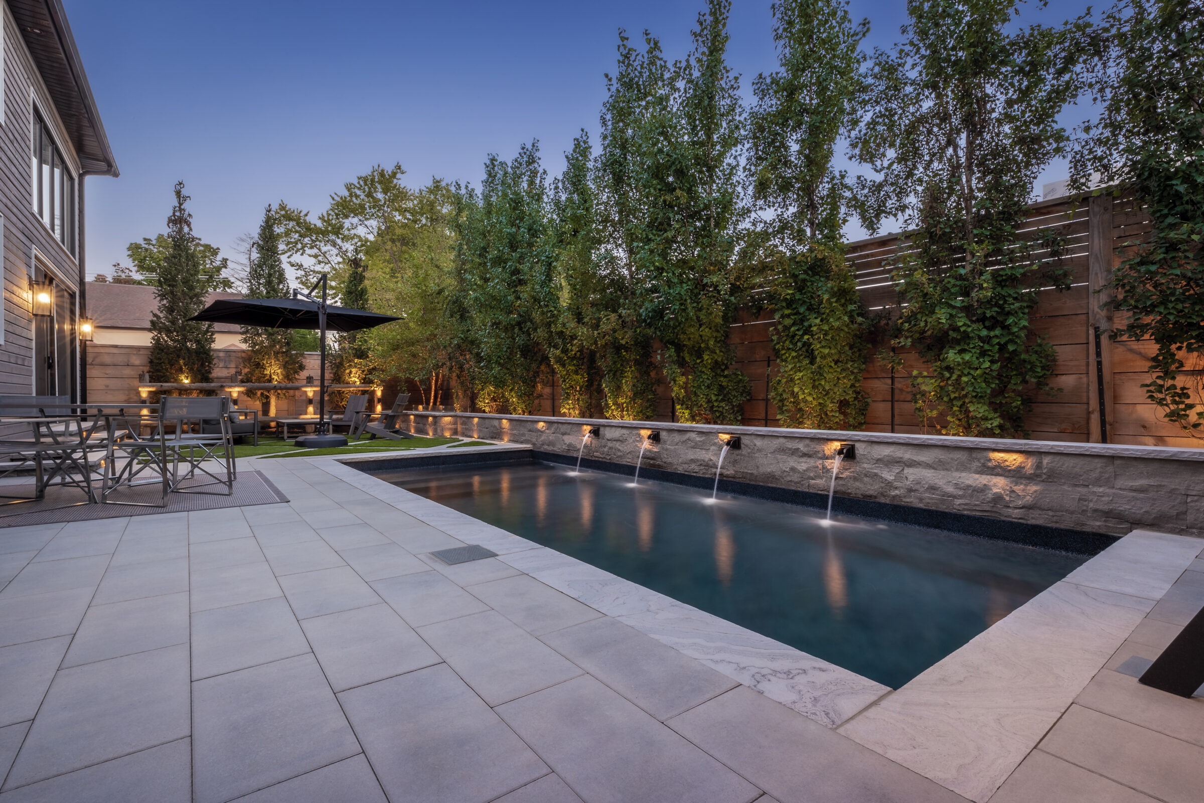 Modern backyard with swimming pool, water features, and patio furniture; surrounded by lush trees and ambient lighting, creating a serene evening atmosphere.
