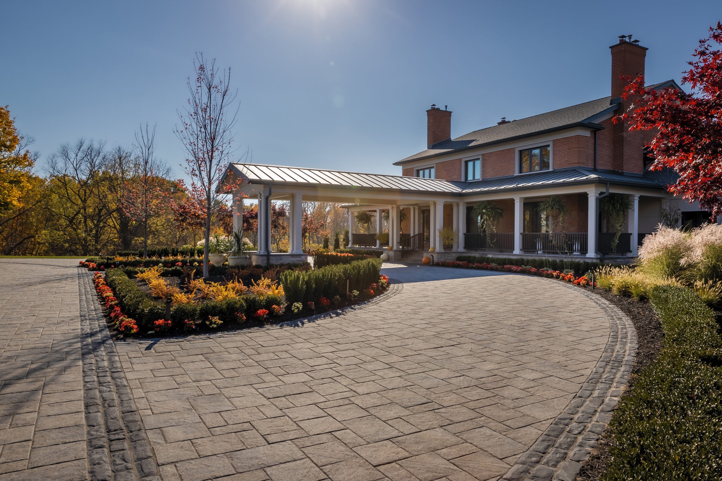 Charming house with brick facade, landscaped garden, and curved driveway under clear sky. Surrounded by autumn foliage, featuring elegant porch and columns.
