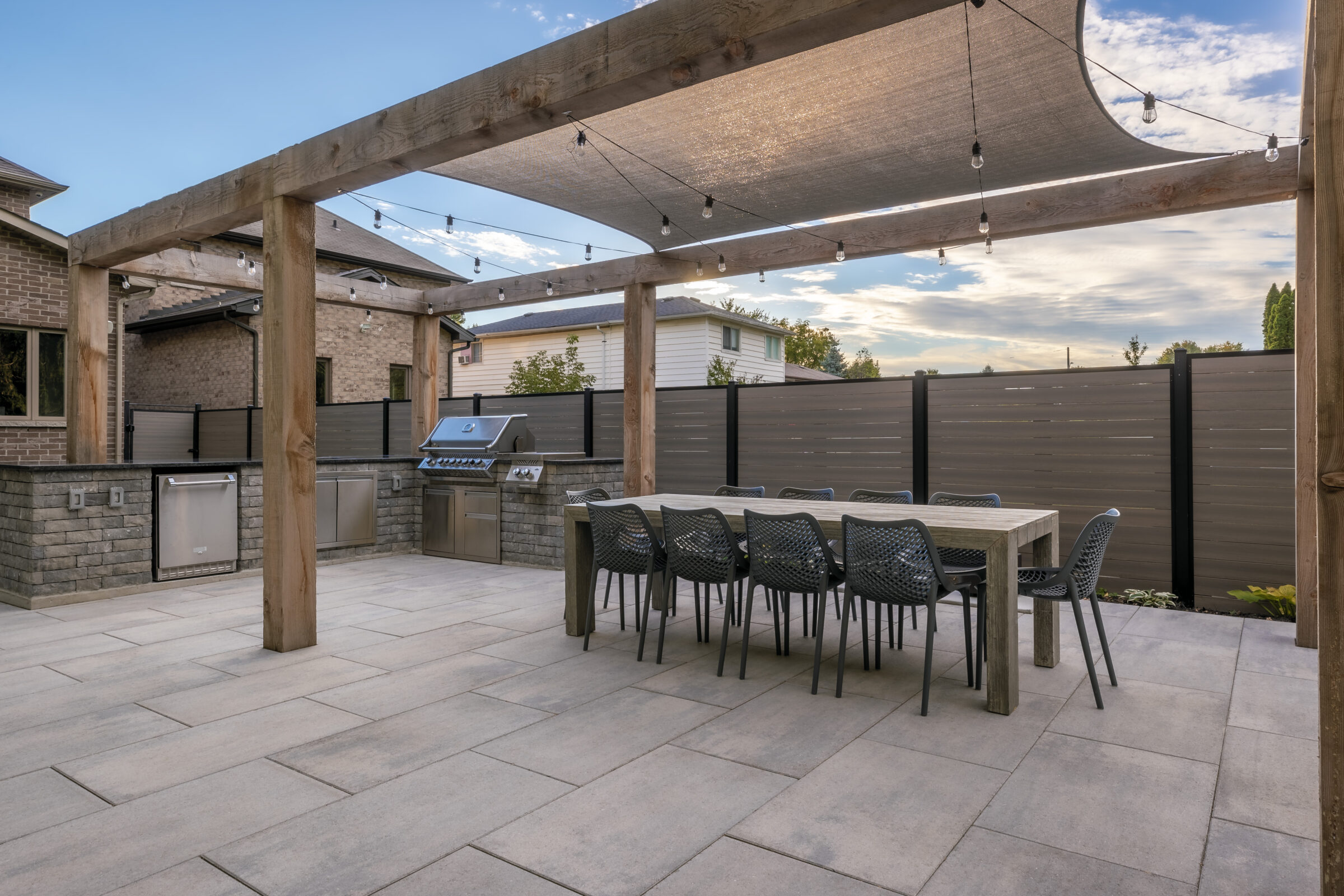 Spacious backyard patio features a large dining table, string lights, pergola, and built-in stainless steel grill against a fenced yard backdrop.