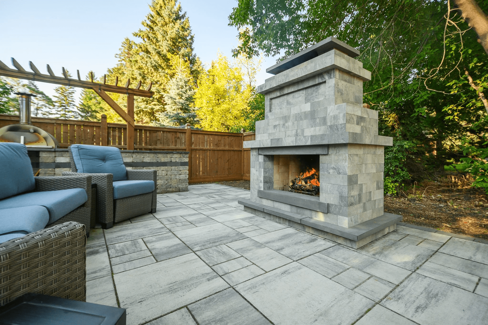 Outdoor patio with gray stone fireplace, wicker seating, and wooden fence. Surrounded by lush trees, creating a cozy, natural atmosphere.