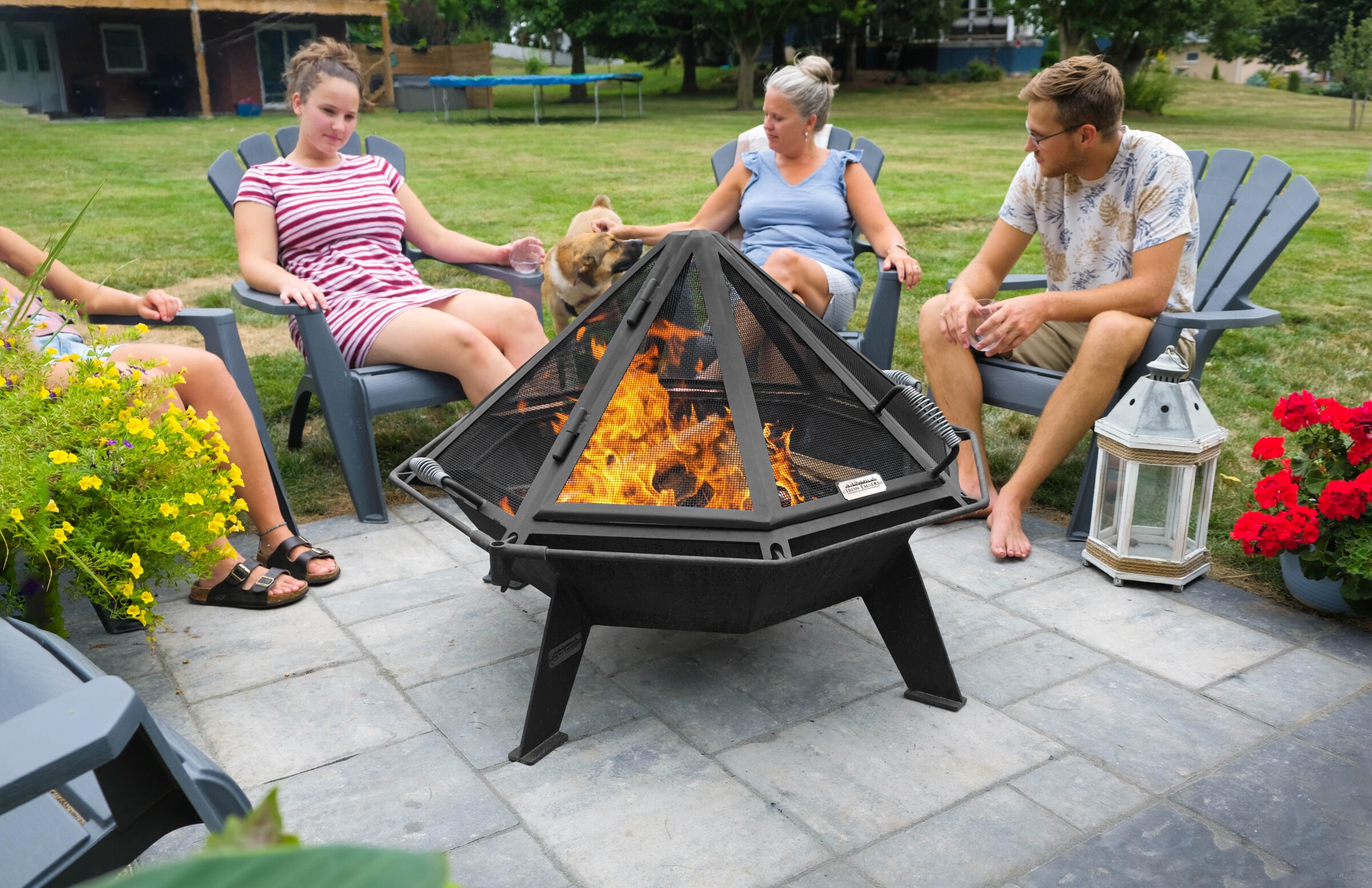 Four people sit around a backyard fire pit with a dog, surrounded by flowers and greenery on a sunny day.