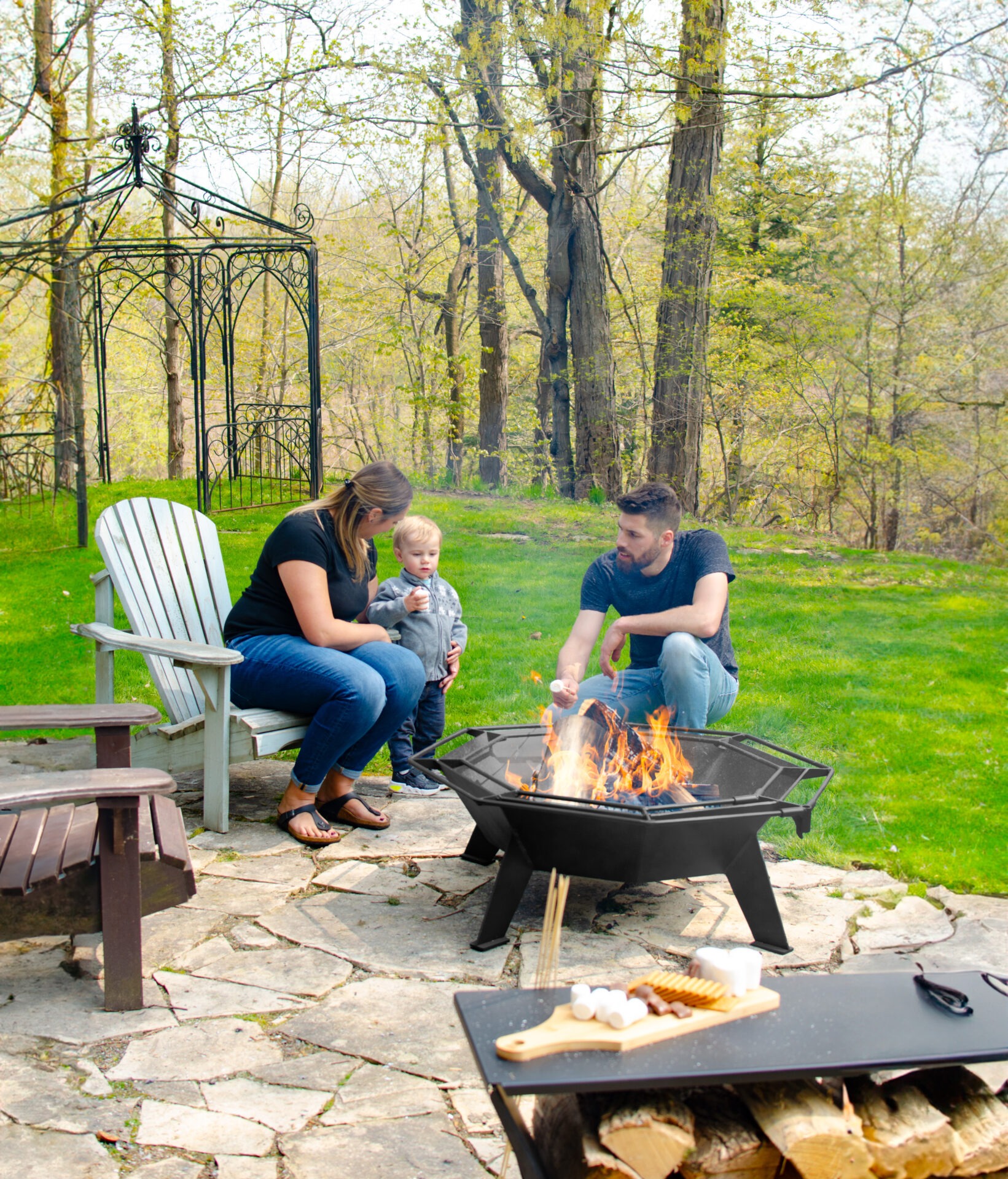 Three people enjoy a backyard campfire, roasting marshmallows amidst trees. Adirondack chairs encircle the fire pit on a stone patio.