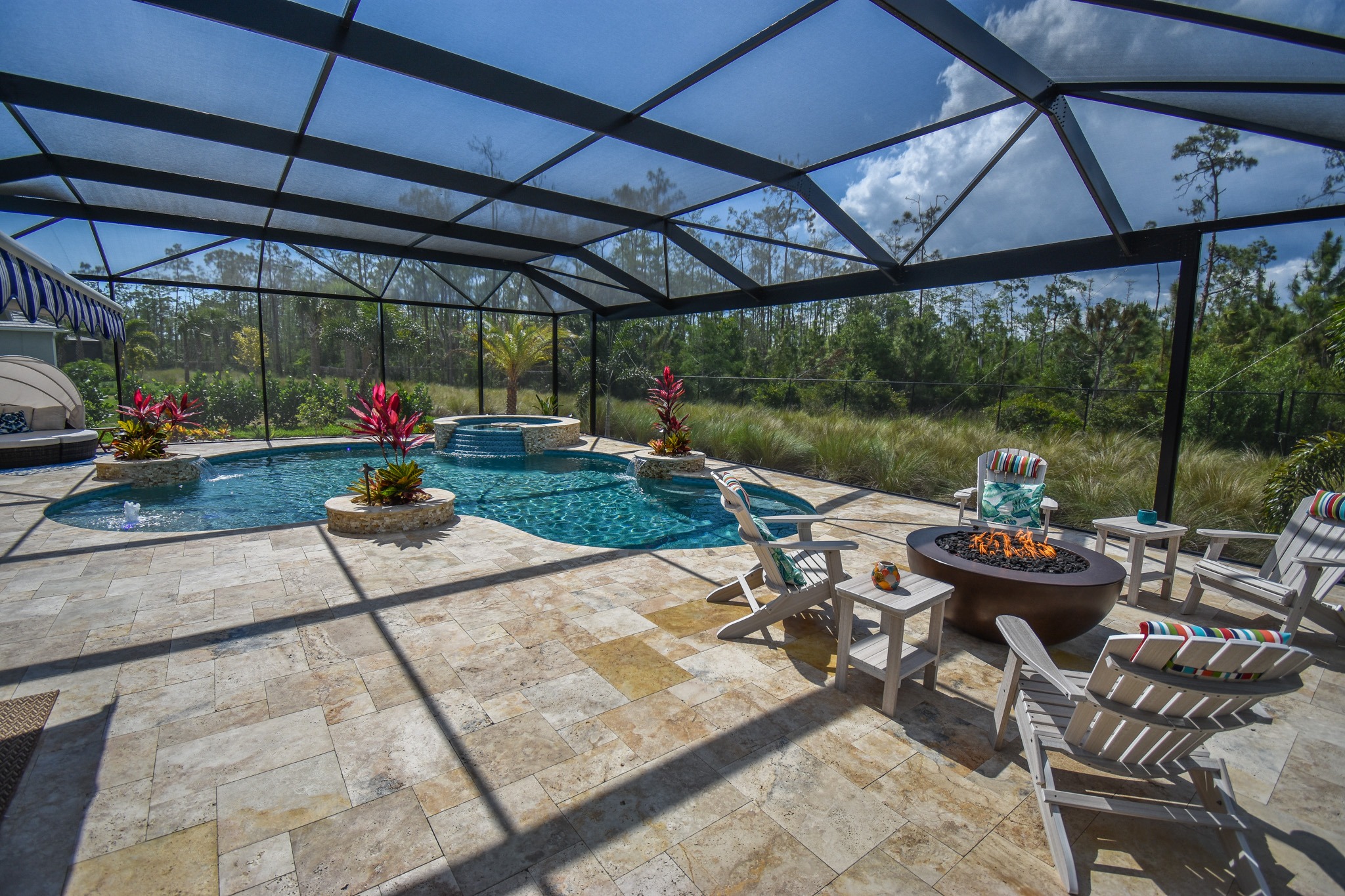 Enclosed patio with a swimming pool, spa, and fire pit surrounded by chairs. Lush greenery outside, under a sunny, cloud-dappled sky.