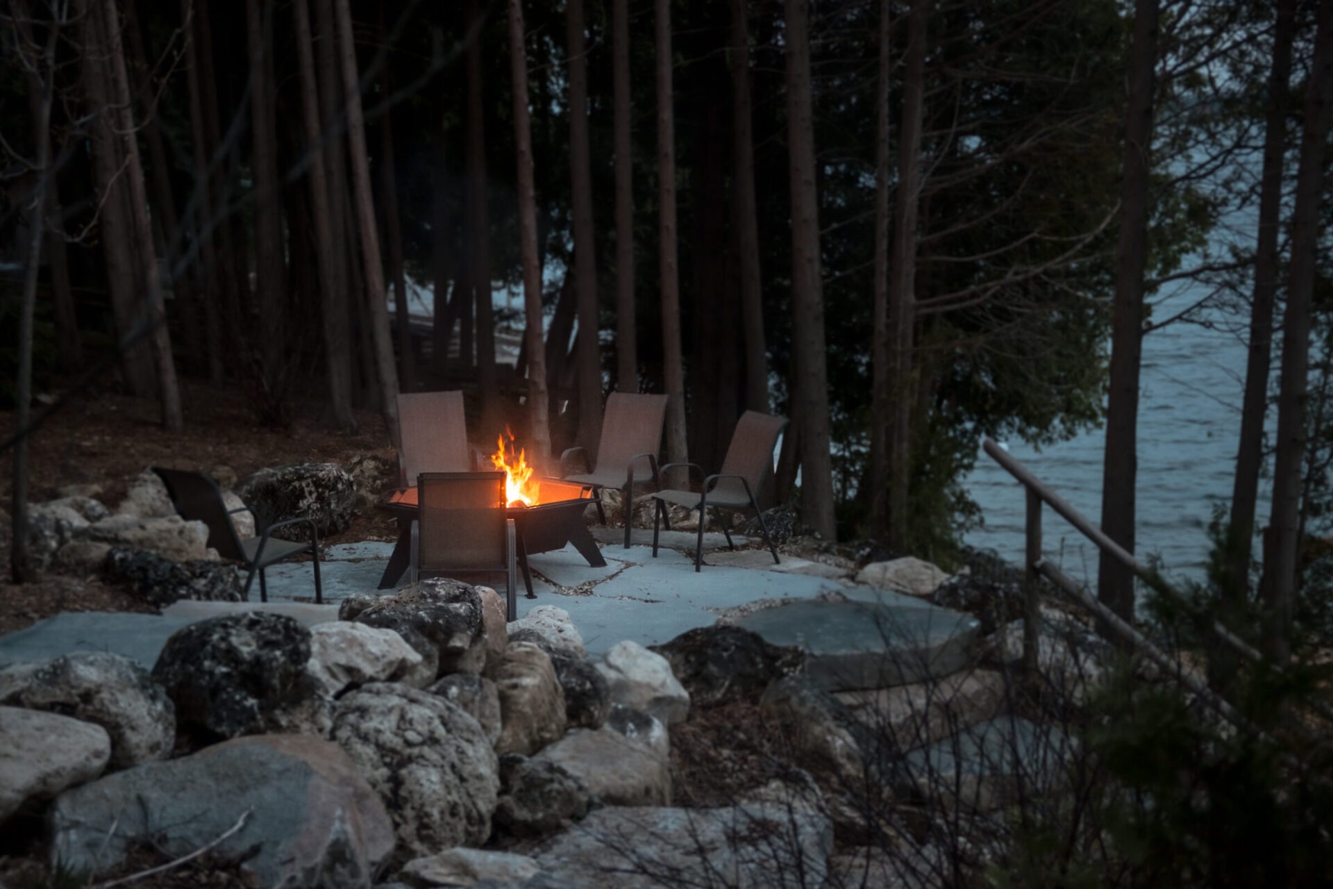 A cozy outdoor fire pit is surrounded by chairs amid trees, overlooking a tranquil lakeside setting with stone pathways and natural ambiance.