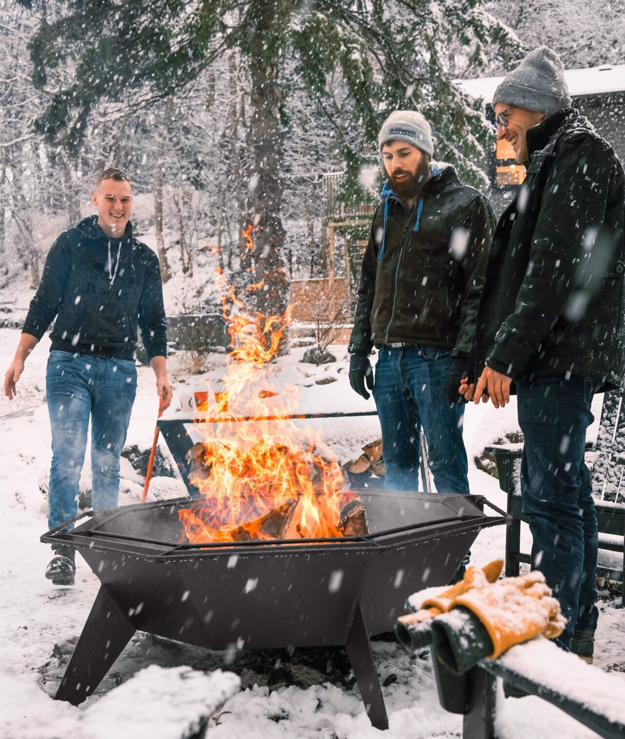 Three people stand around a large fire pit in a snowy, forested setting, staying warm amidst falling snowflakes.