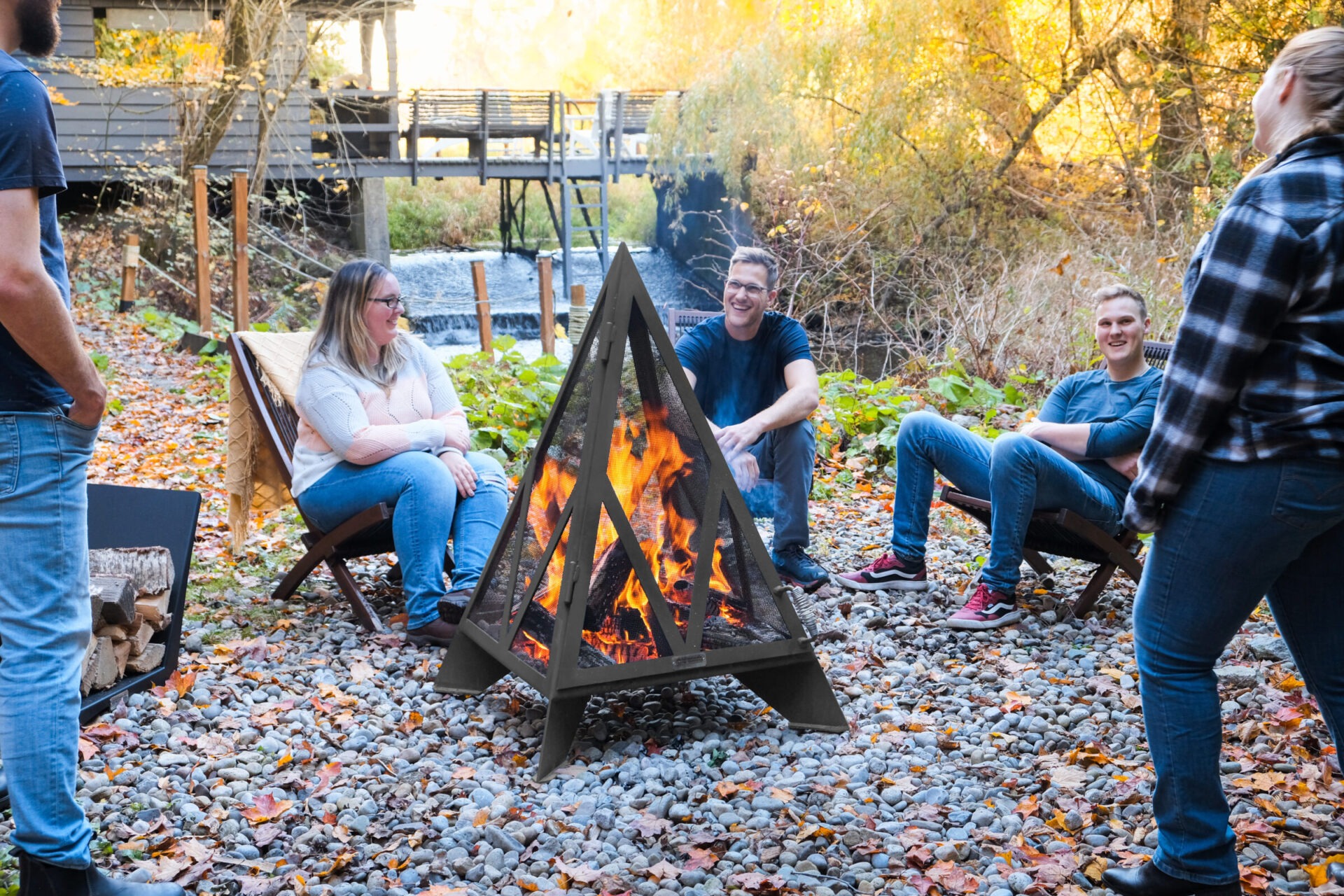 Four people sit around a campfire in a wooded area, surrounded by autumn leaves and a wooden bridge nearby.