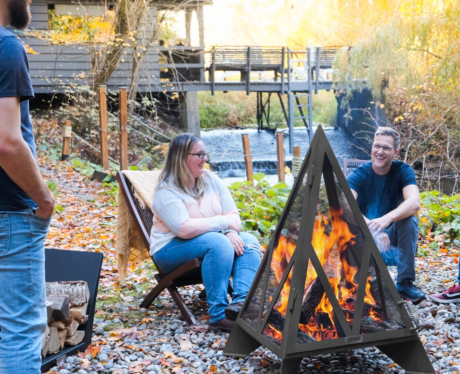 Four people sit around a campfire in a wooded area, surrounded by autumn leaves and a wooden bridge nearby.