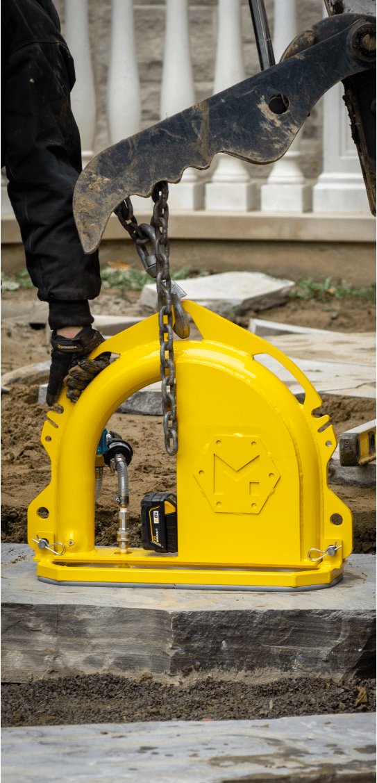 A construction site with a person operating machinery lifting a bright yellow device, surrounded by stone slabs and soil.