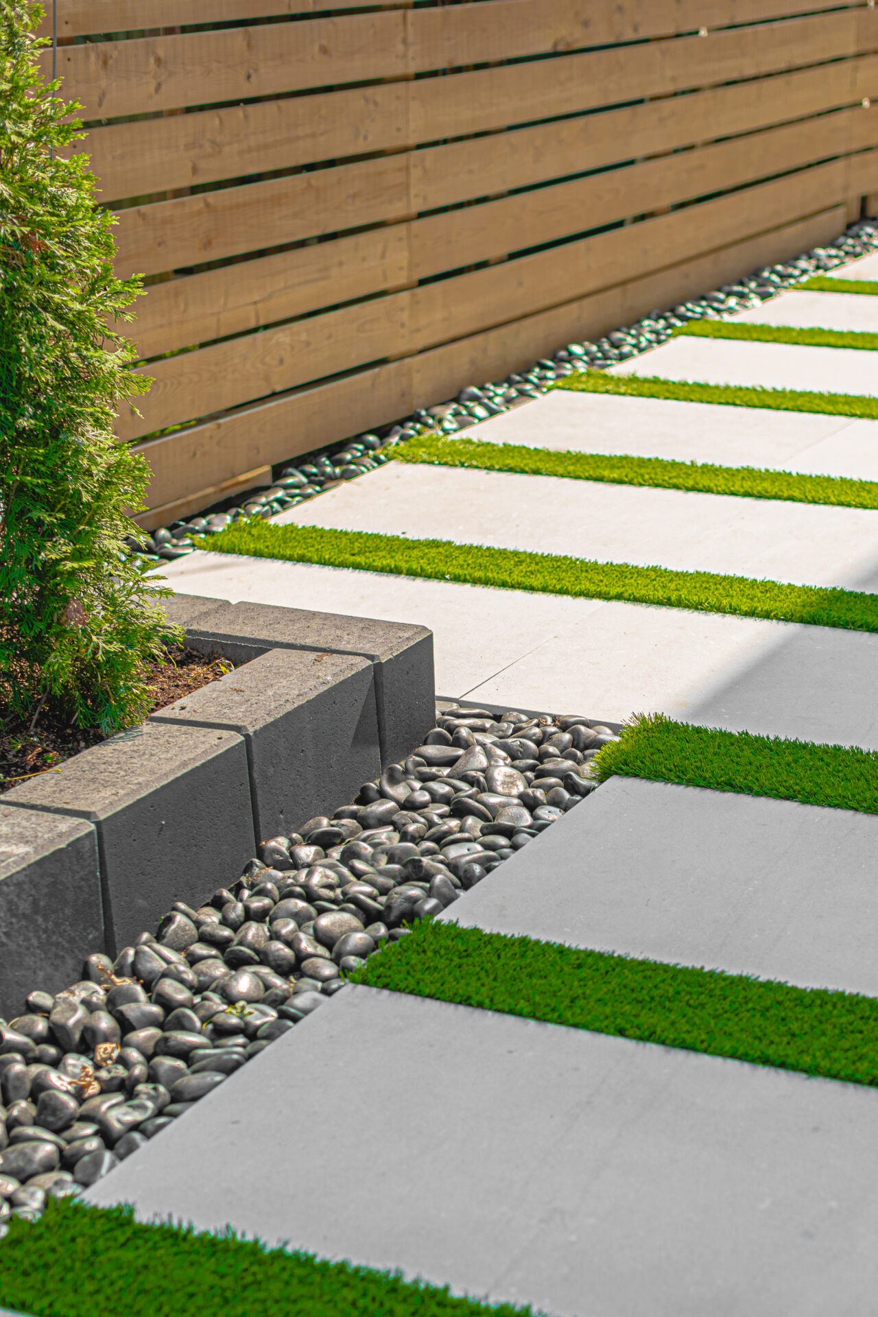 Modern garden path with rectangular concrete slabs, artificial grass strips, and smooth stones. Adjacent to a wooden fence and evergreen shrub.
