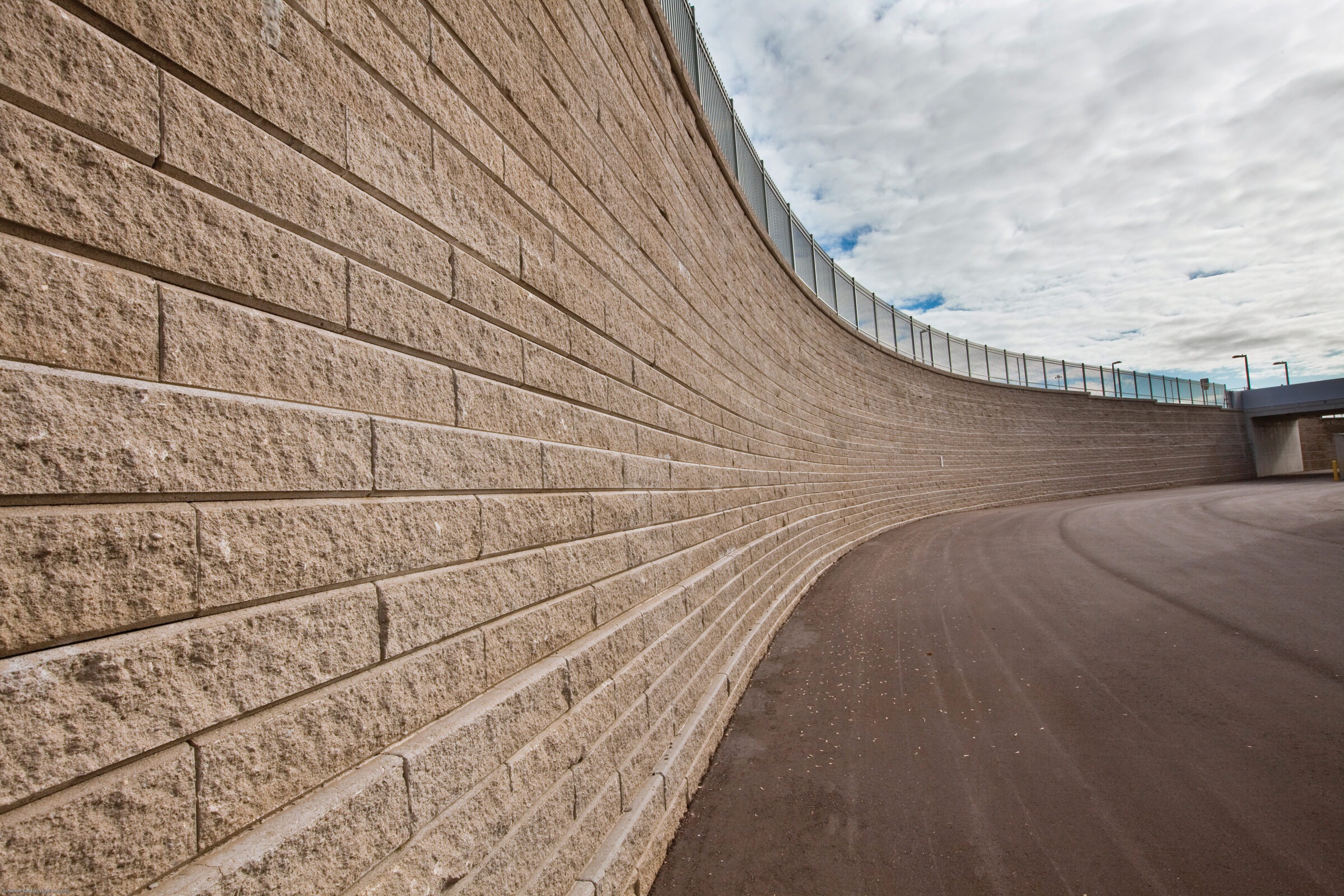 Curved, textured stone retaining wall with metal fencing above, adjacent to a smooth asphalt pathway under a cloudy blue sky.