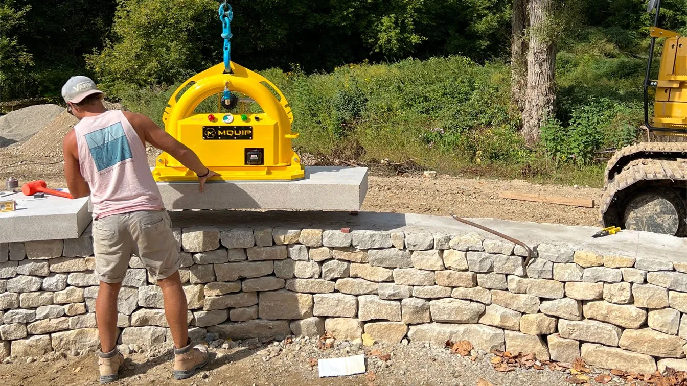 A person operates a yellow lifting machine to place concrete slabs on a stone wall at a construction site.