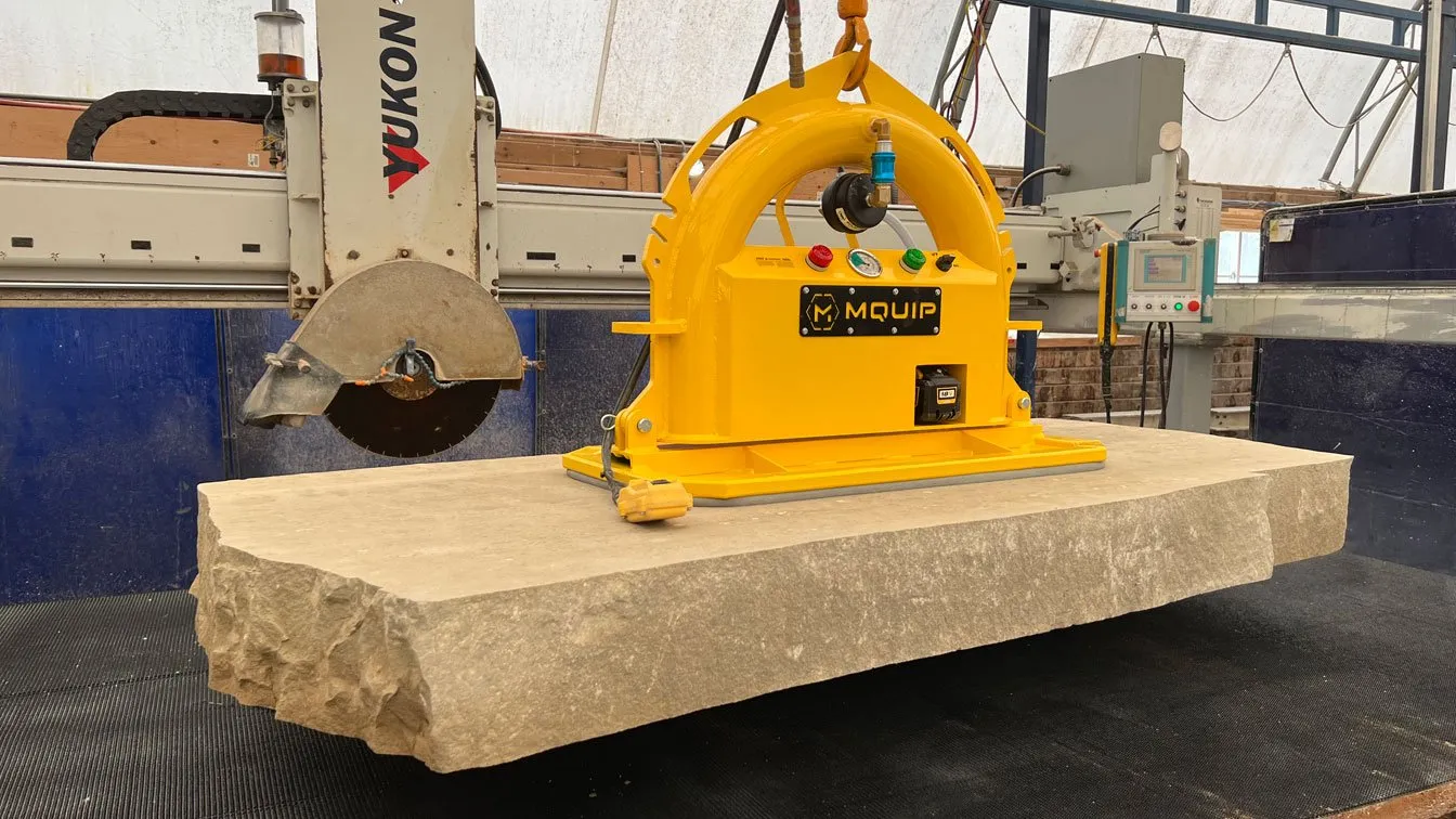 A large yellow machine hoists a hefty stone slab beneath a cutting saw in an industrial workshop setting.