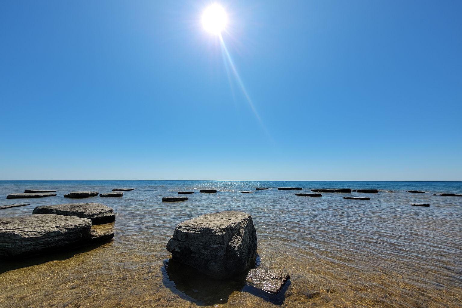 The image shows a tranquil seascape under a bright sun, with flat rocks scattered across the clear, shallow water.