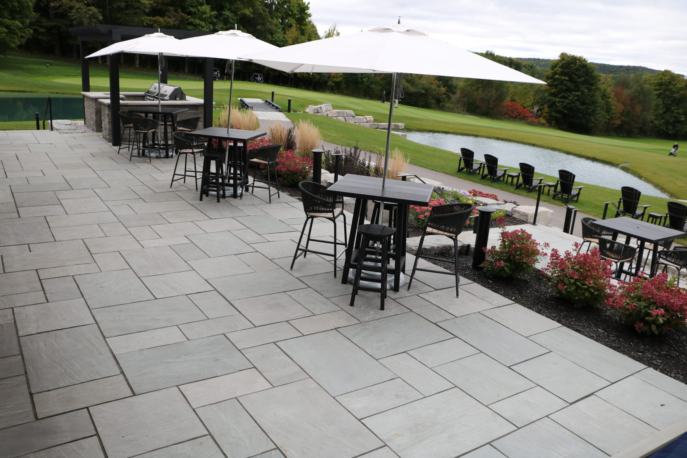 Spacious outdoor patio with tables, chairs, and umbrellas overlooking a serene pond, surrounded by lush greenery and blooming flowers, under a cloudy sky.