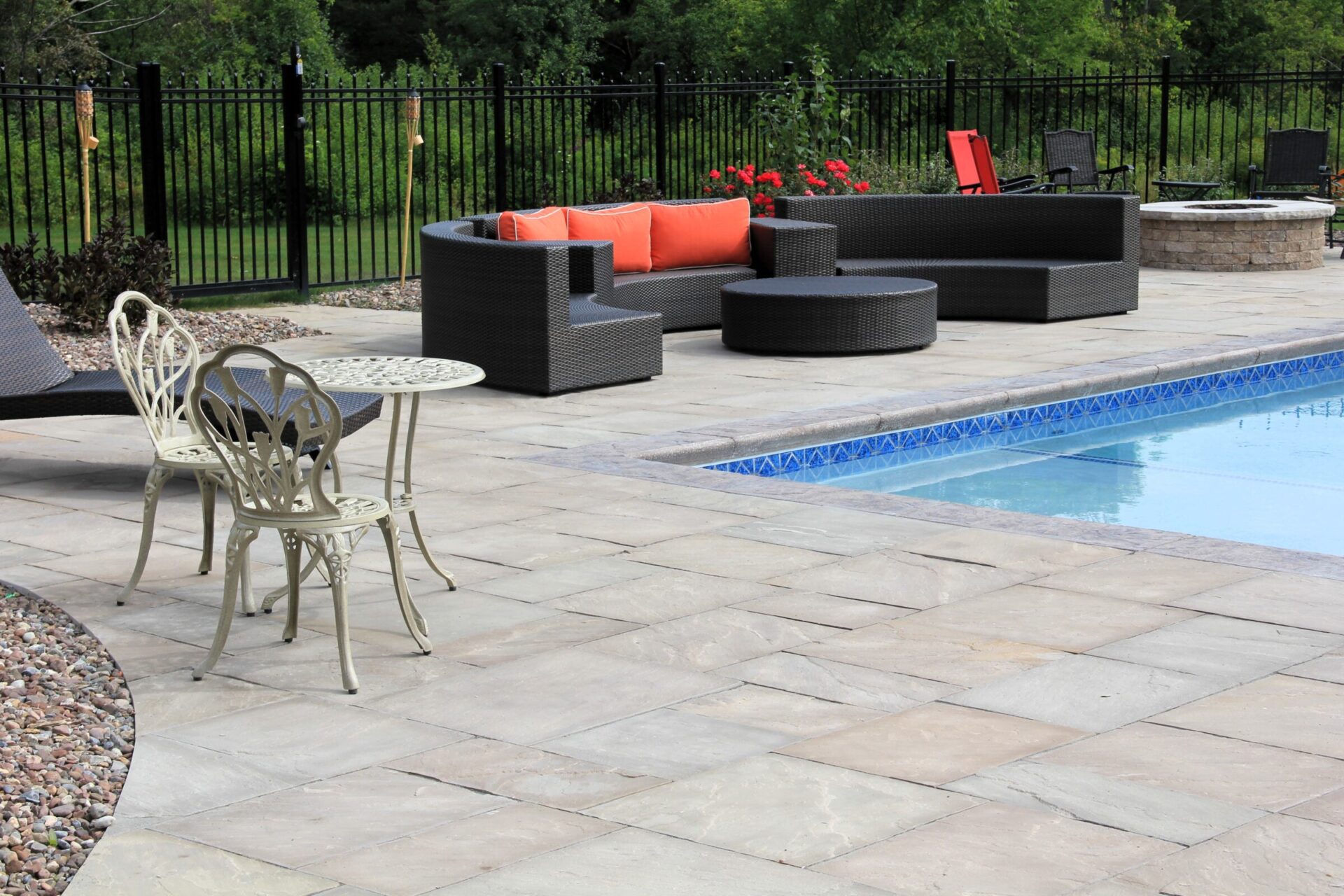 Spacious poolside patio with cushioned wicker furniture, bistro table, and lush greenery beyond a black metal fence. Relaxed ambiance for outdoor gatherings.