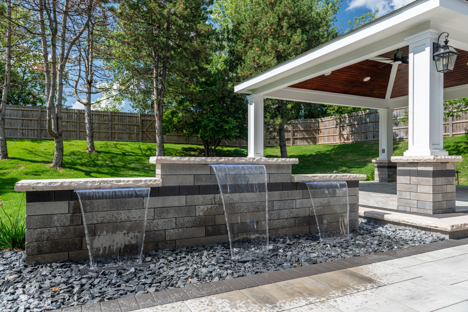 A modern outdoor area features a covered patio and cascading water feature, surrounded by trees and greenery, under a clear blue sky.