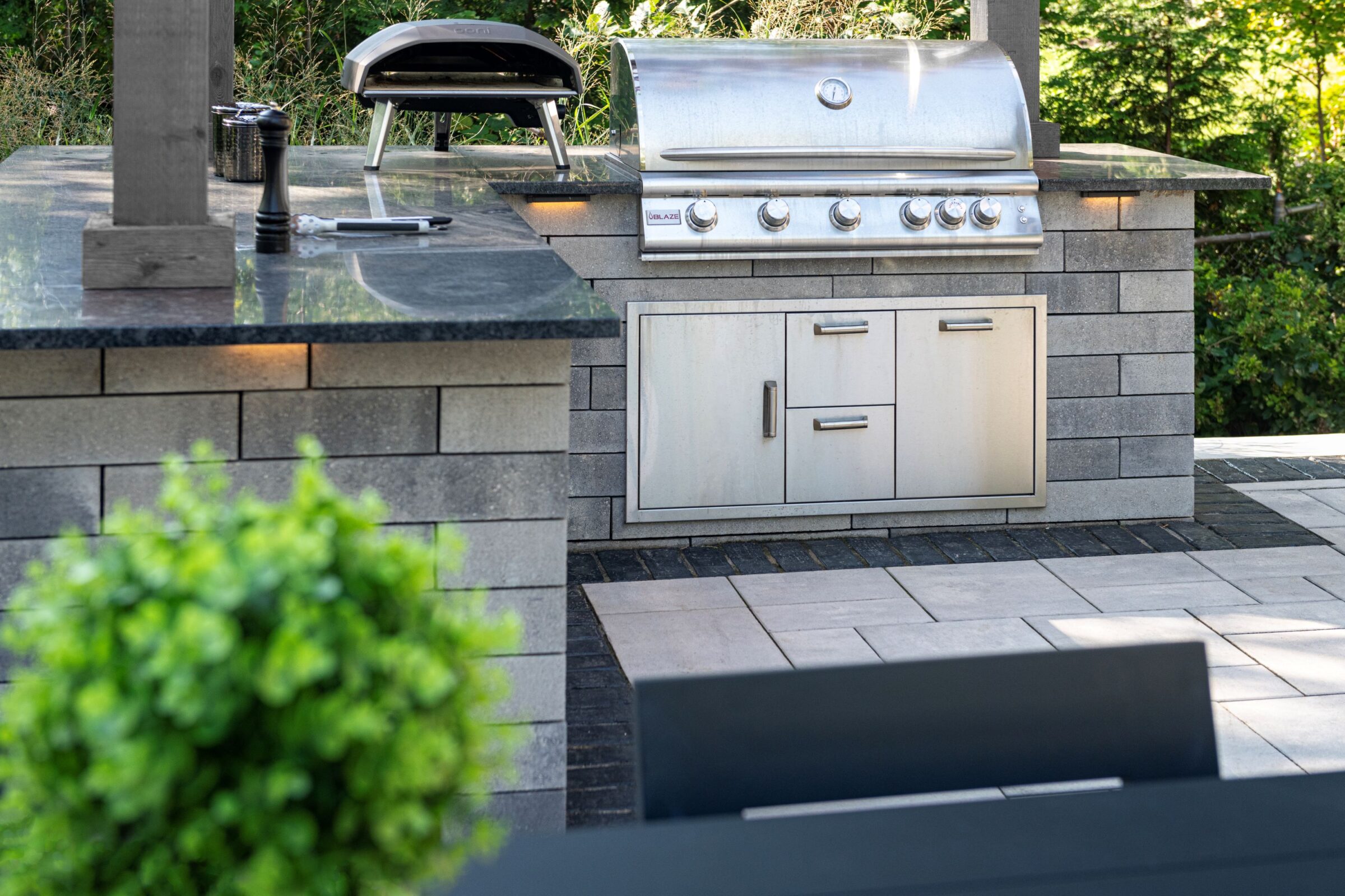 Outdoor kitchen with stainless steel grill, pizza oven, and modern stone countertop, surrounded by greenery and tiled patio flooring.