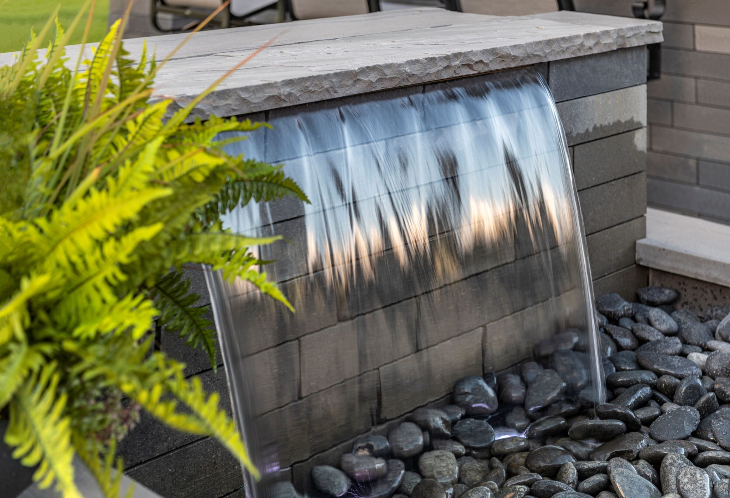 A small waterfall flows over a stone ledge onto smooth black pebbles, with green ferns adding natural elegance to the scene.