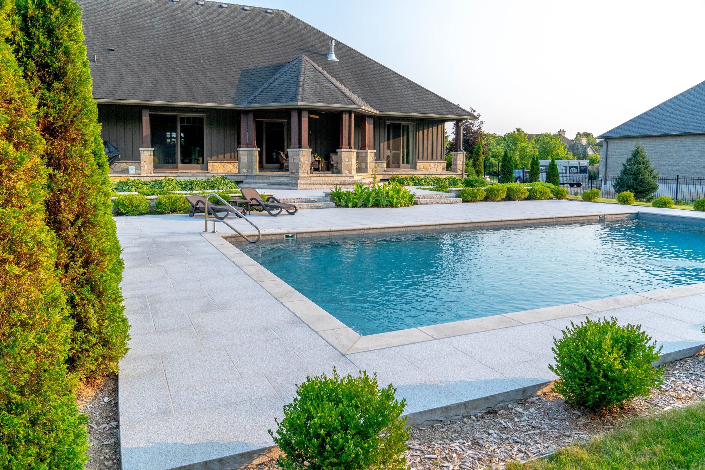 Spacious backyard with a swimming pool, patio area, and chairs. Lush greenery surrounds a large house. Peaceful, modern outdoor setting.