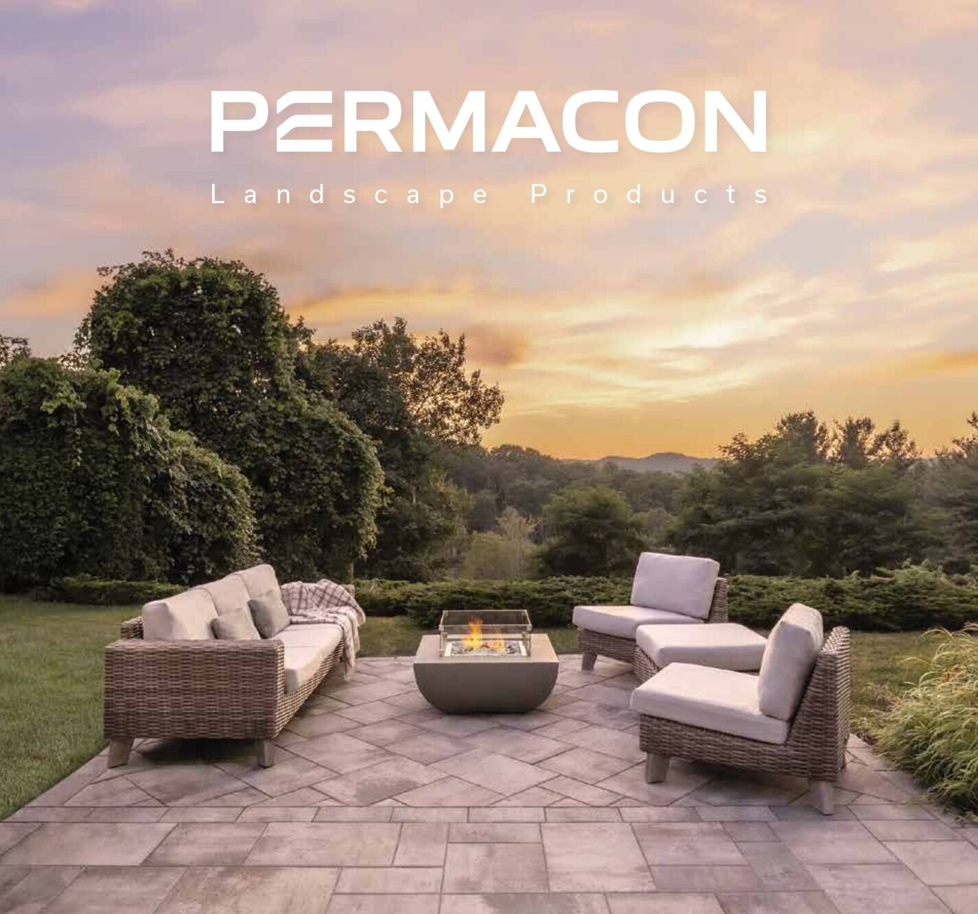 Patio setup with wicker furniture and fire pit overlooking a scenic, wooded landscape at sunset. Text reads “Permacon Landscape Products.”