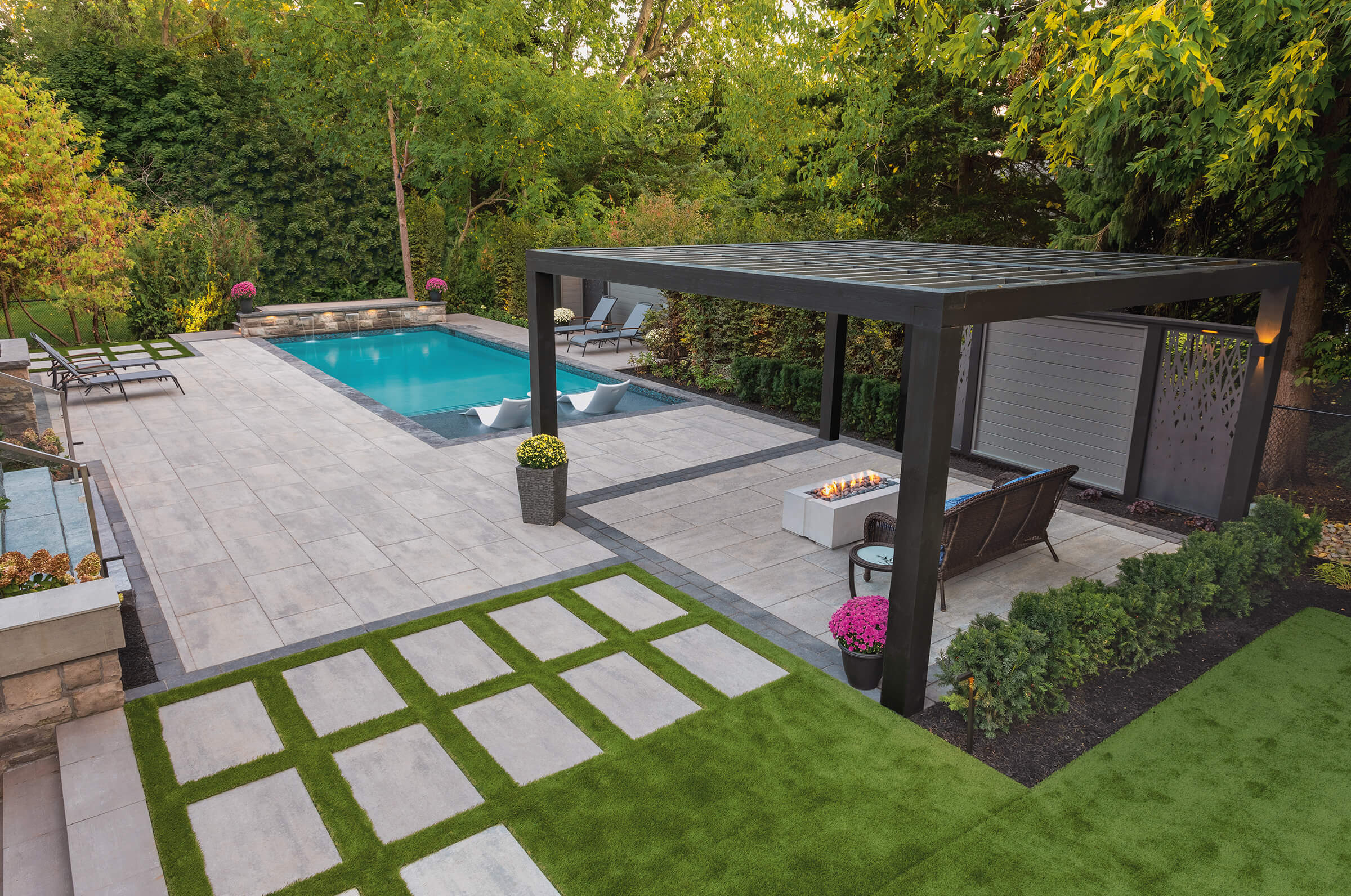 Modern backyard features a pool, pergola, seating area, fire pit, and decorative plants. Green lawn contrasts with stone tiles, surrounded by trees.