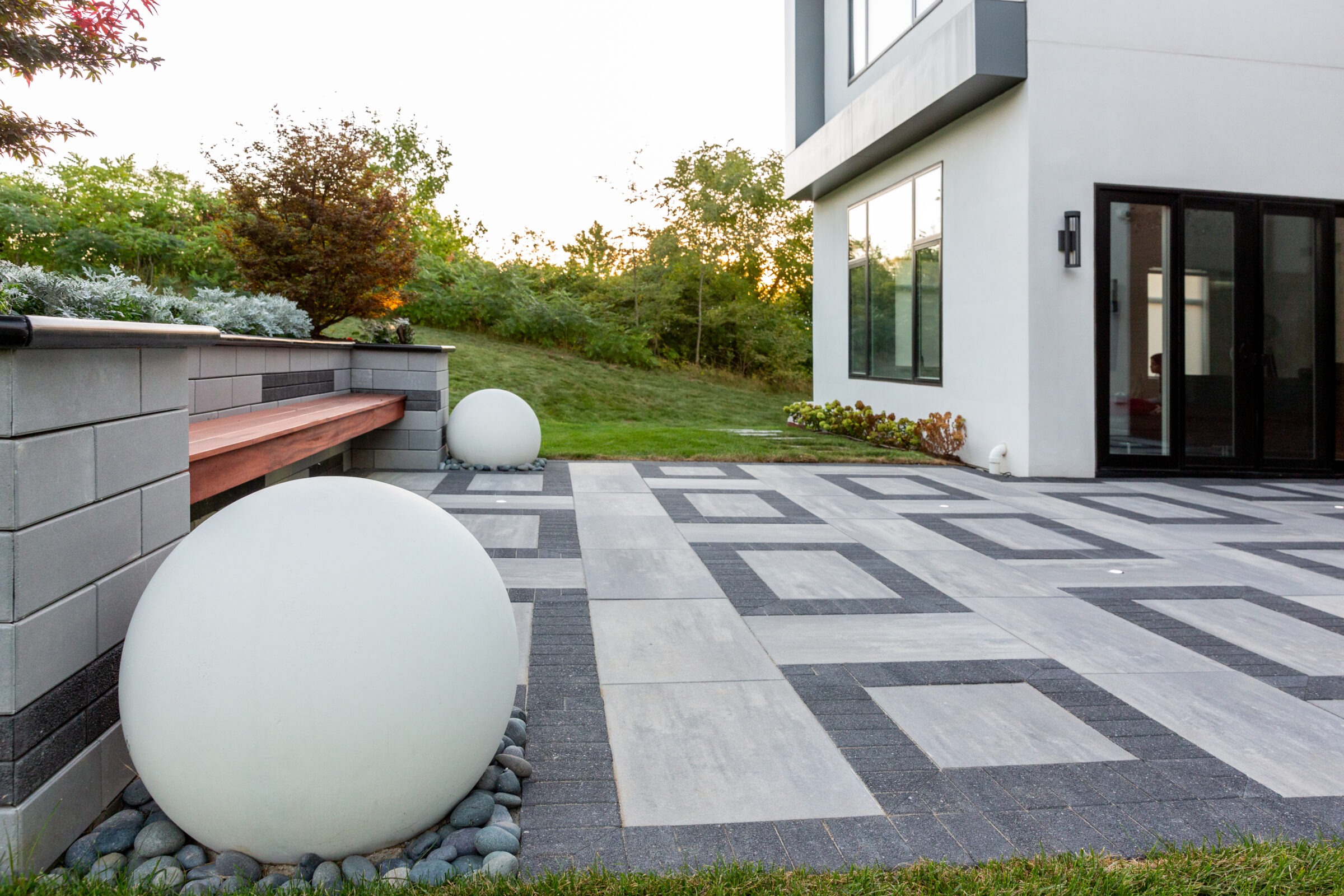 Modern patio with geometric paving, white spheres, and minimalist landscaping. Adjacent to a contemporary building with large windows and sliding glass doors.