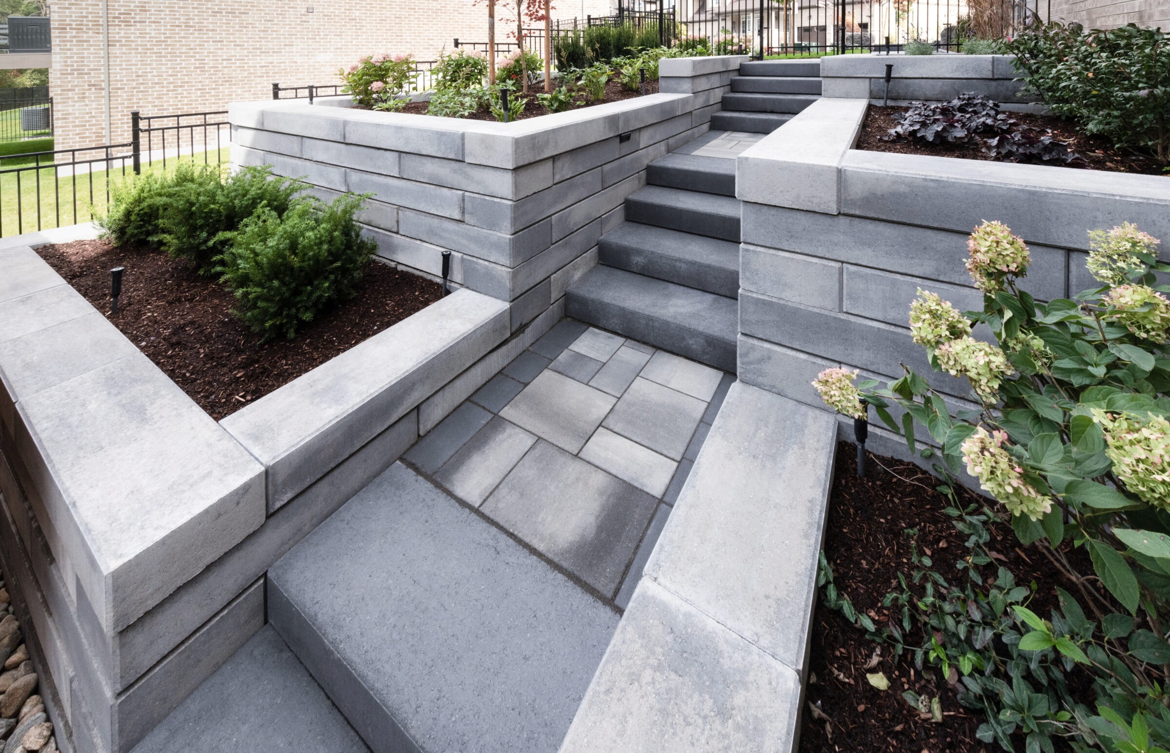 Modern garden with terraced stone walls, stairs, and neatly arranged plants. Bright and tidy outdoor design adjacent to a brick building.