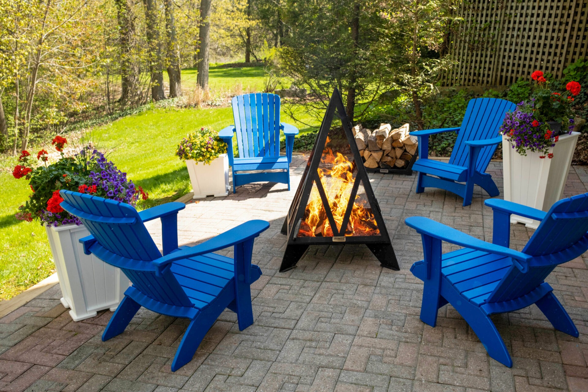 Blue Adirondack chairs surround a modern hexagonal fire pit on a patio, with colorful flowers and lush green backyard scenery.