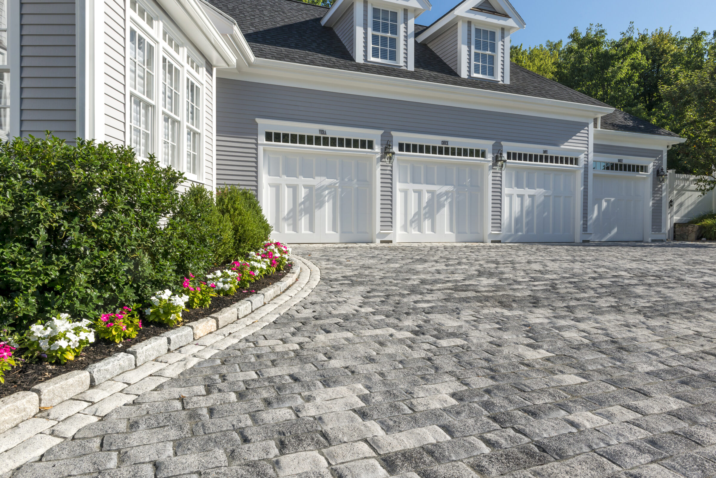 A large garage with three white doors and dormer windows, bordered by a cobblestone driveway and colorful flower beds.