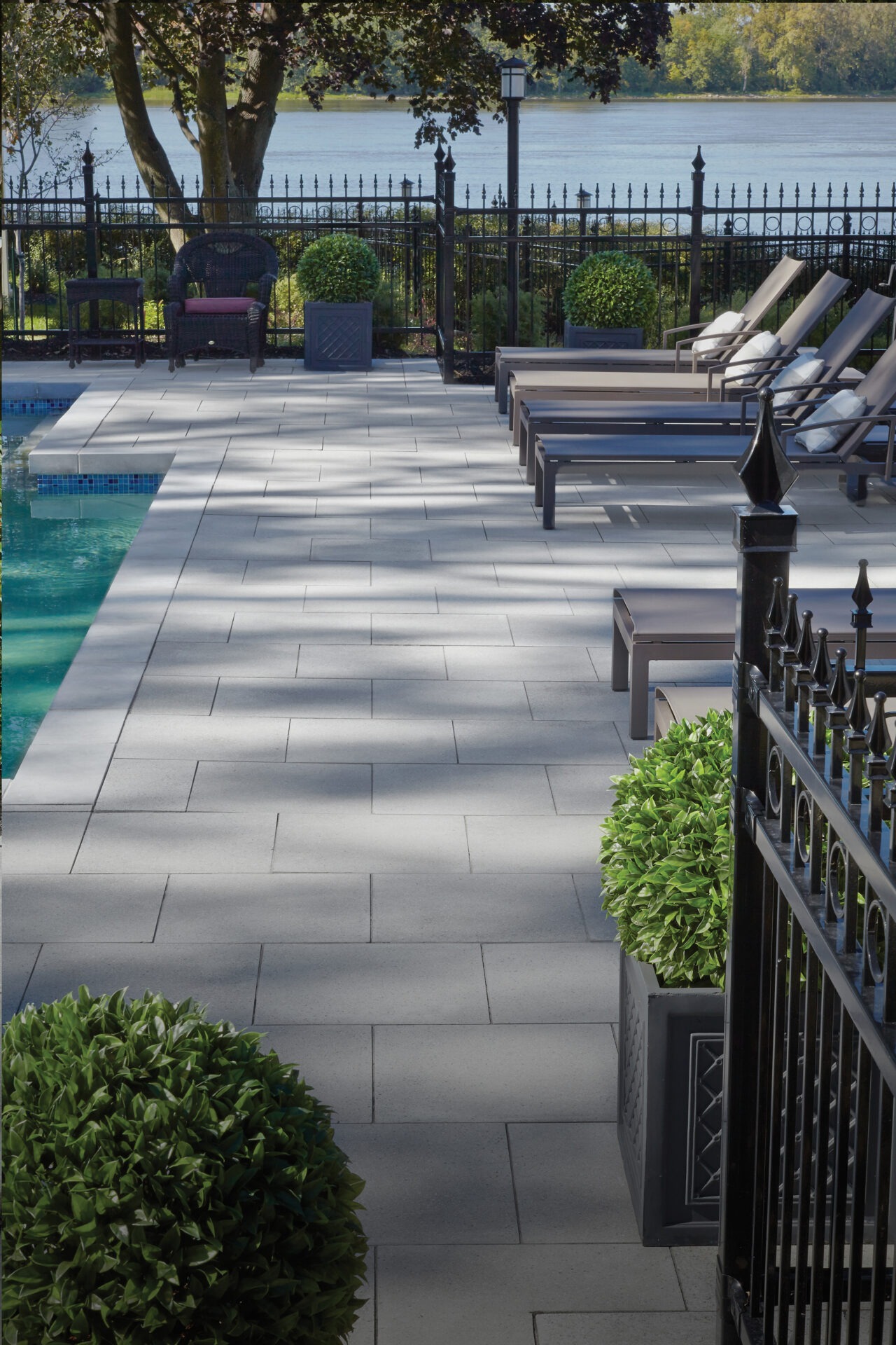 Poolside scene with lounge chairs, potted plants, and a view of a calm river beyond a decorative fence. Shadows create a dappled effect.