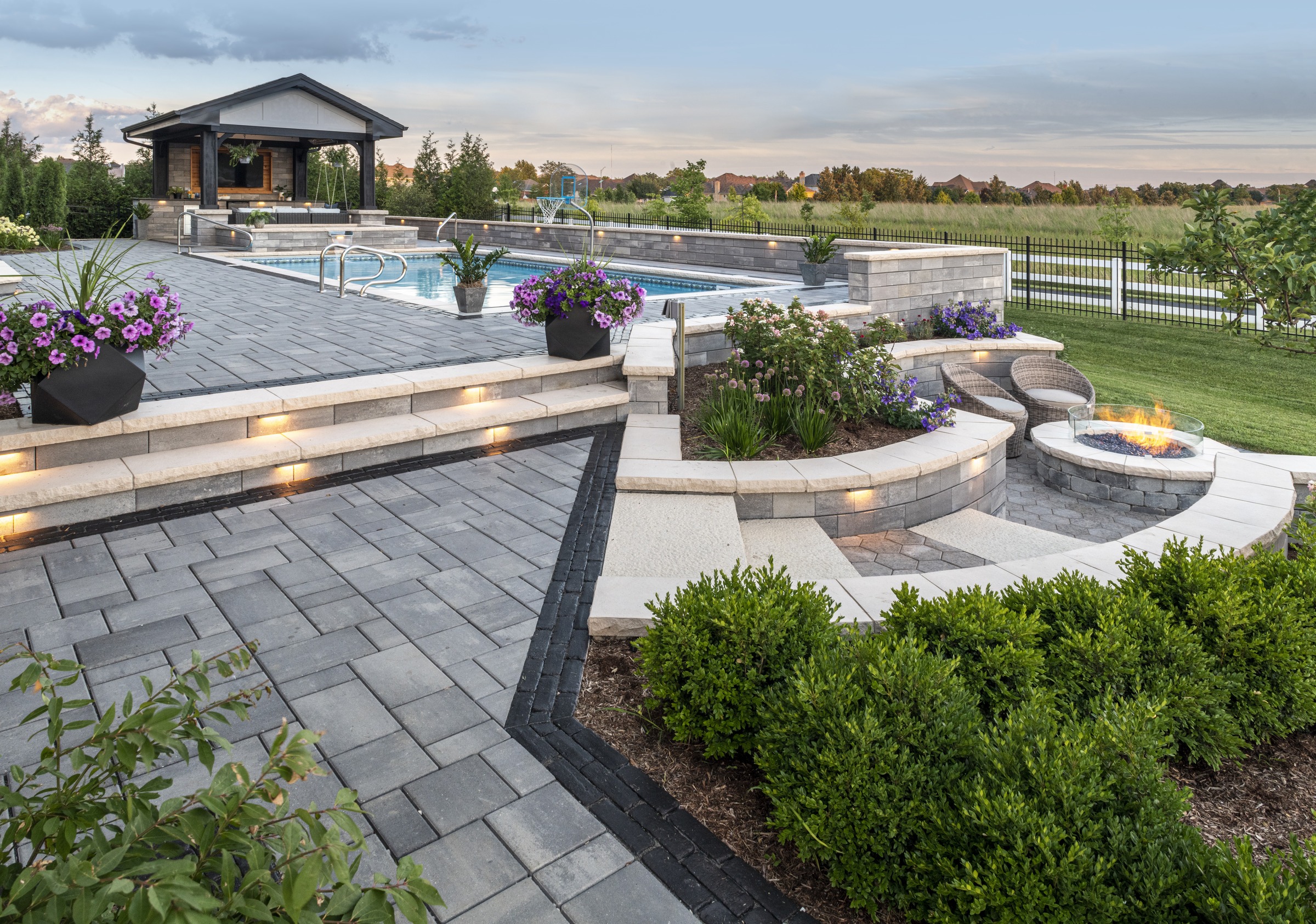 Modern backyard with pool, fire pit, and landscaped garden. Stone patio, steps, colorful flowers, and seating area under a cloudy sky.