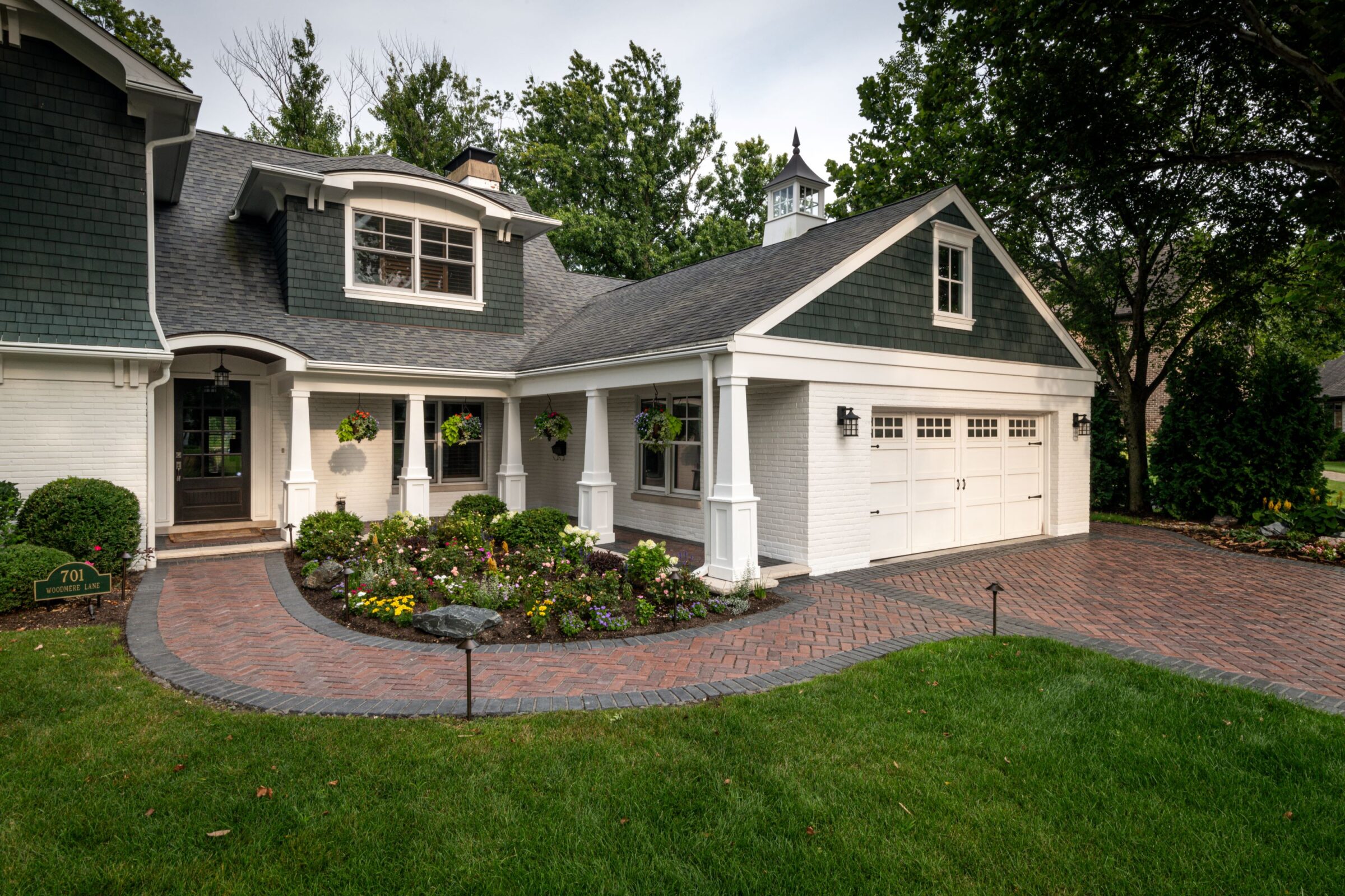 Charming two-story house with a brick driveway, green and white exterior, manicured garden, and hanging plants. Peaceful suburban setting with trees.