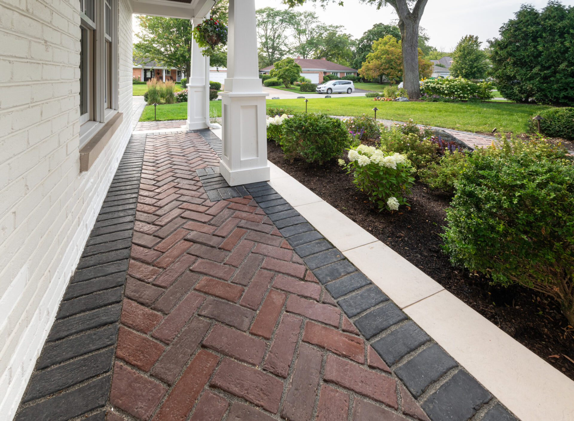 A brick walkway leads to a porch with white columns, surrounded by manicured greenery, and a quiet suburban street with parked vehicle.