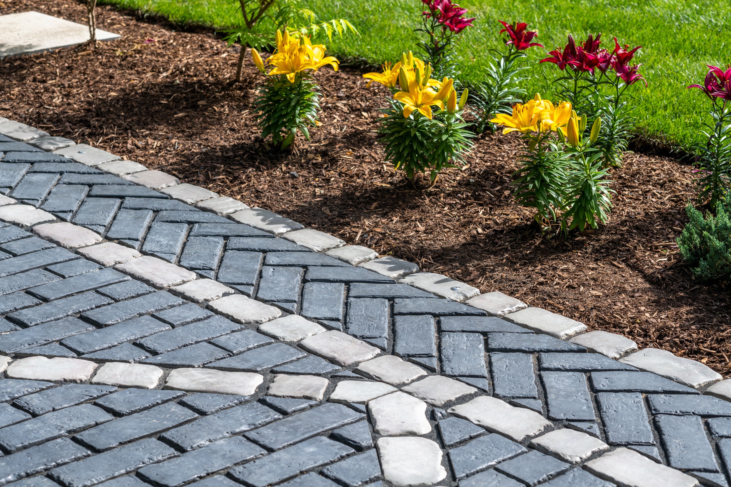 Intricate stone pathway beside vibrant yellow and red flowers, bordered by mulch and lush green grass, creates a serene garden setting.