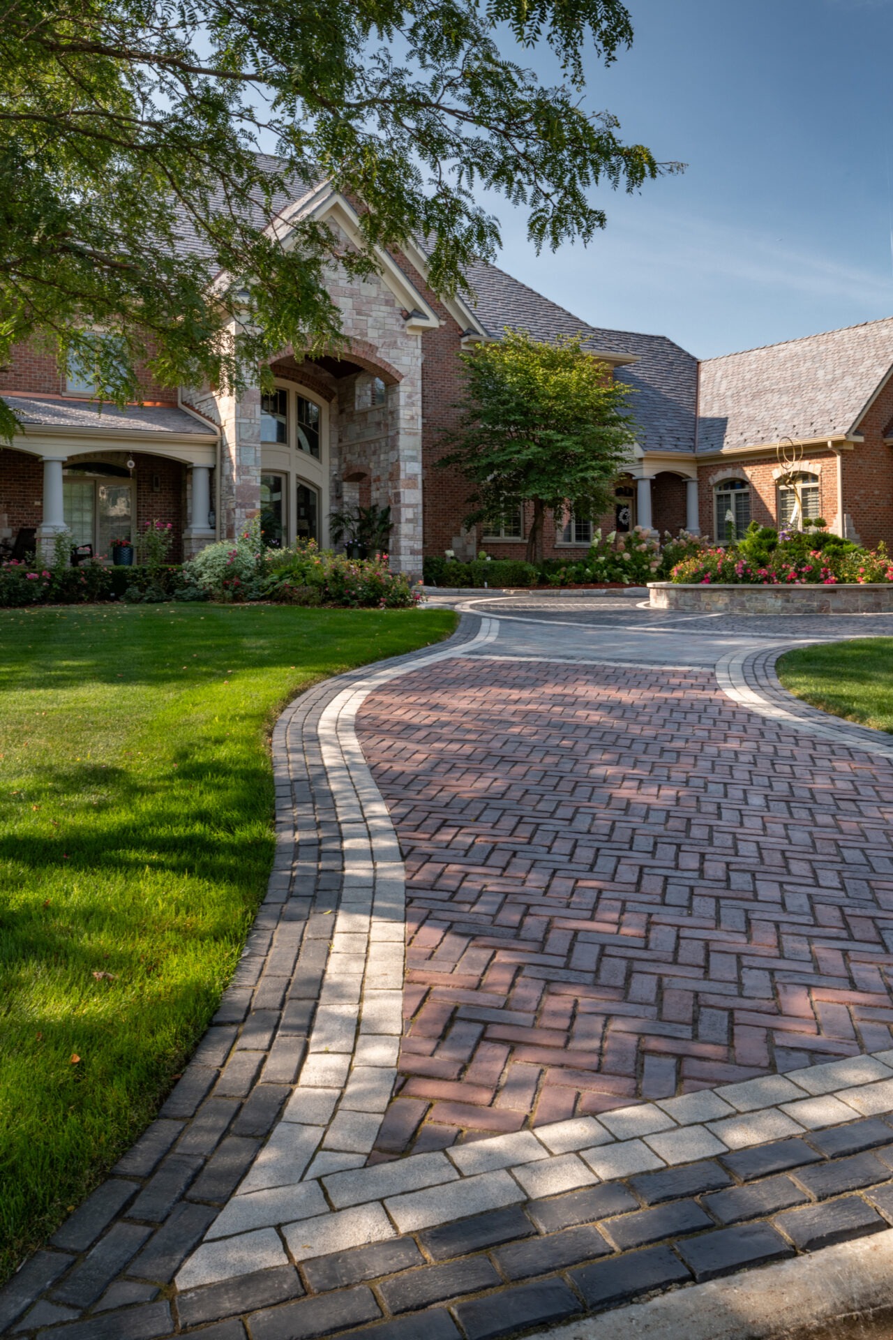 A large brick house with manicured gardens, featuring a winding paved driveway, under a clear blue sky and surrounded by lush greenery.
