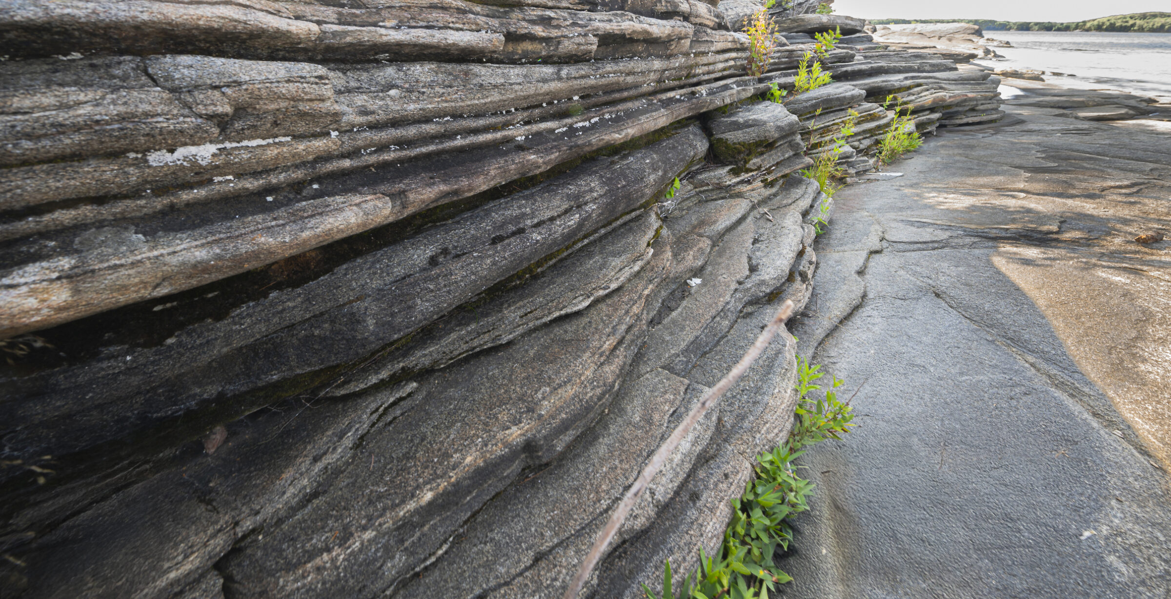 Layered rock formations with green vegetation by a calm riverbank under clear skies, highlighting natural erosion and geological features.