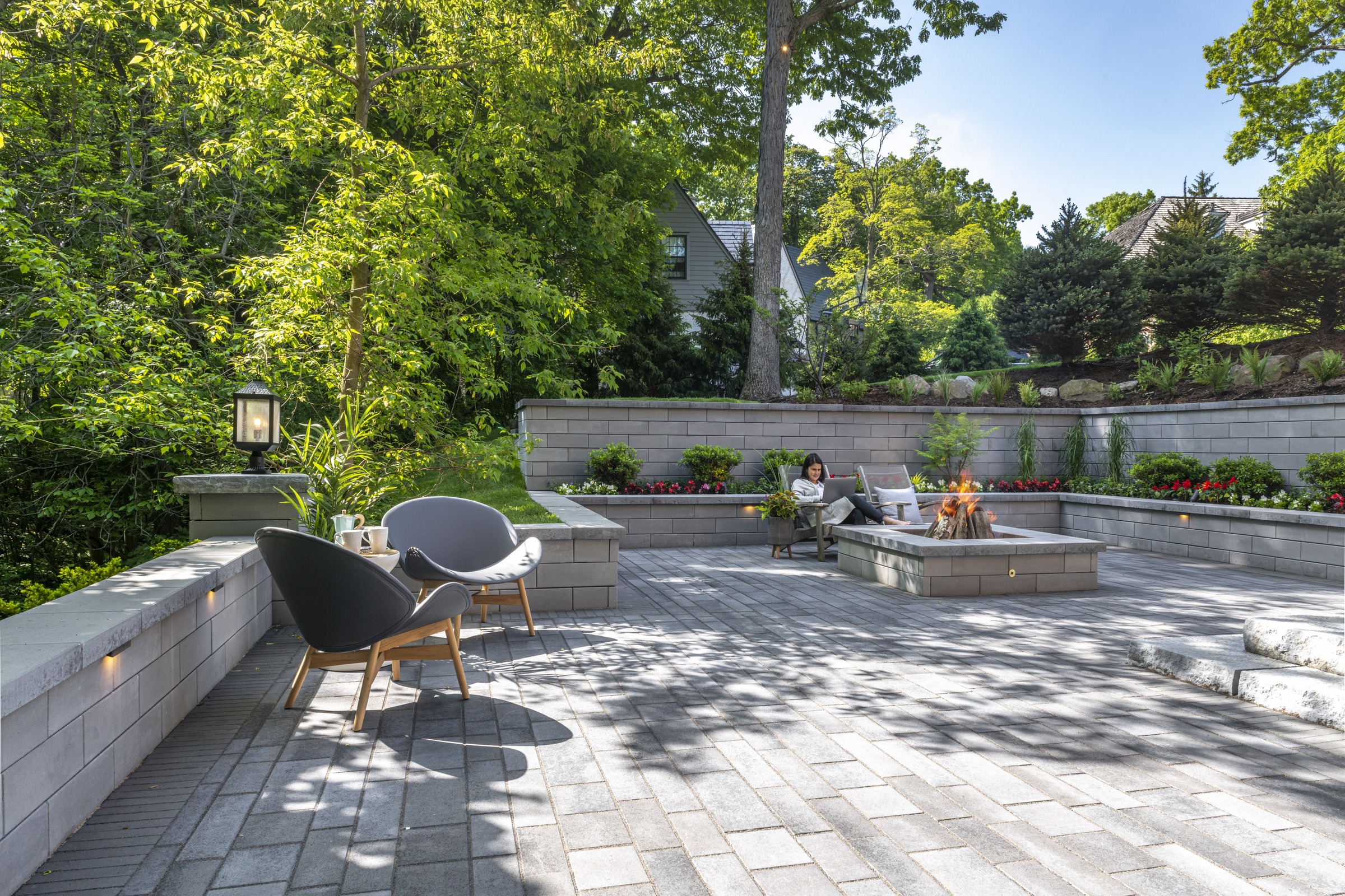 A serene backyard patio features gray seating, a fire pit, and lush greenery. A person relaxes, enjoying the peaceful outdoor setting.
