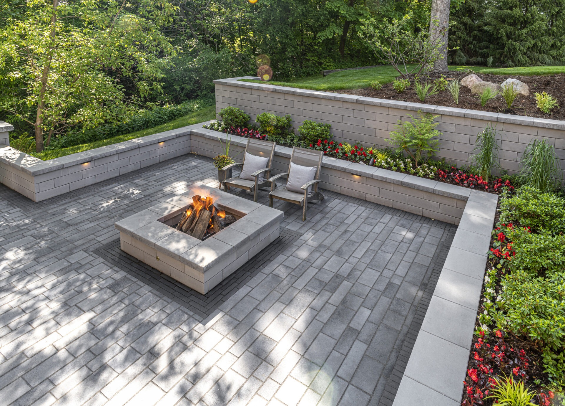 Cozy outdoor patio with a modern fire pit, surrounded by lush greenery and vibrant flowers, featuring two seating chairs on gray pavers.