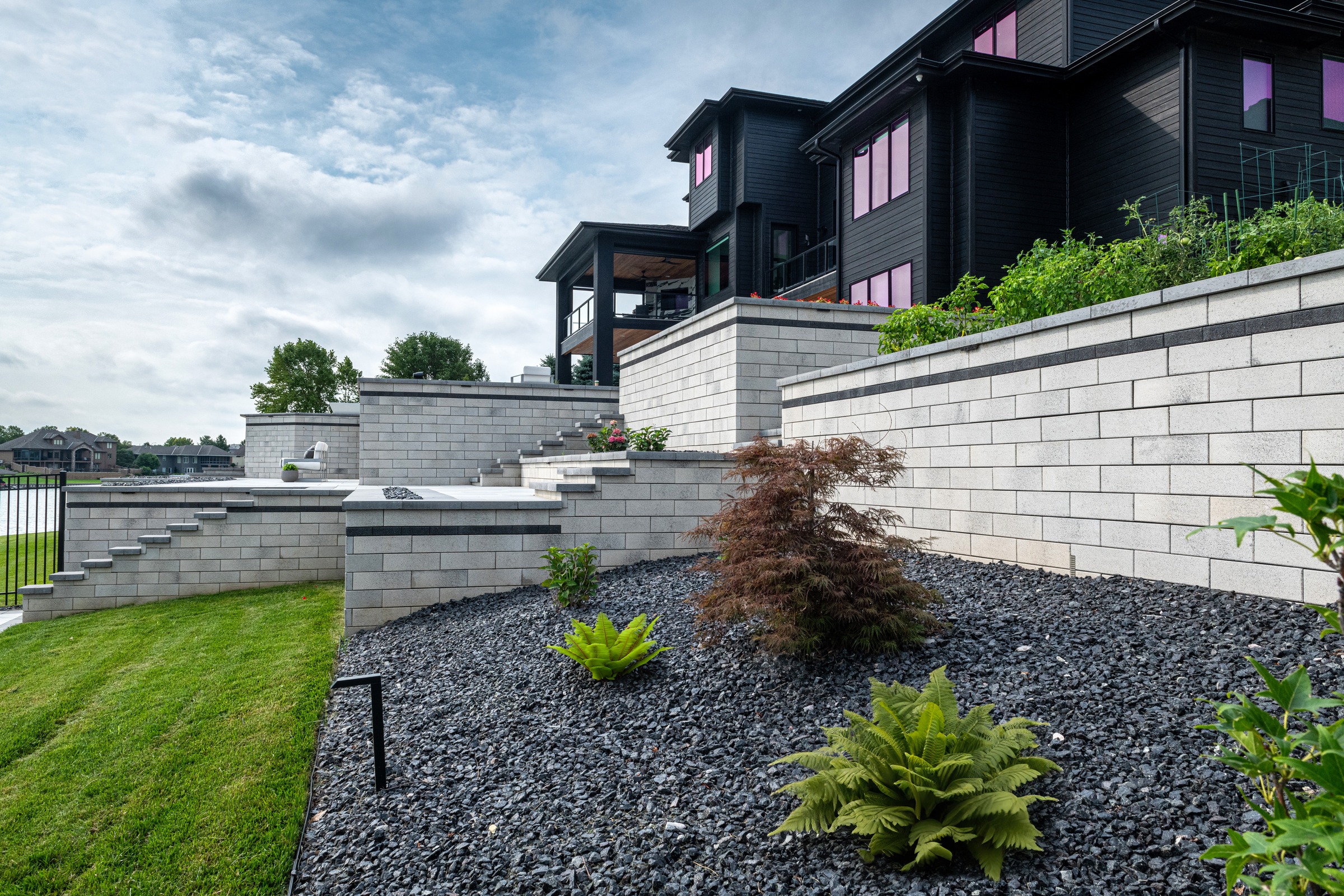 Contemporary black house with terraced stone walls, landscaped with plants, overlooks a lush lawn and a distant lake under a cloudy sky.