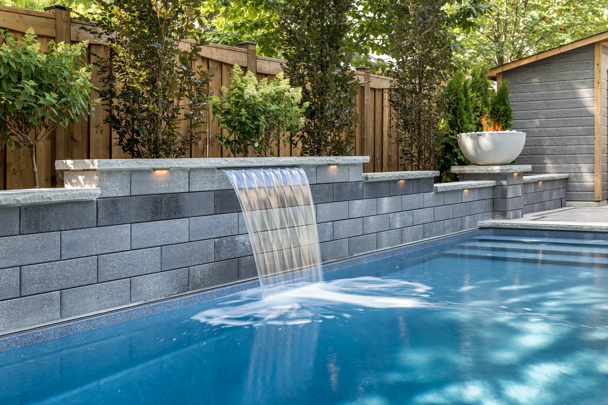 A serene backyard pool features a cascading waterfall, surrounded by lush plants and a wooden fence, creating an inviting, tranquil atmosphere.