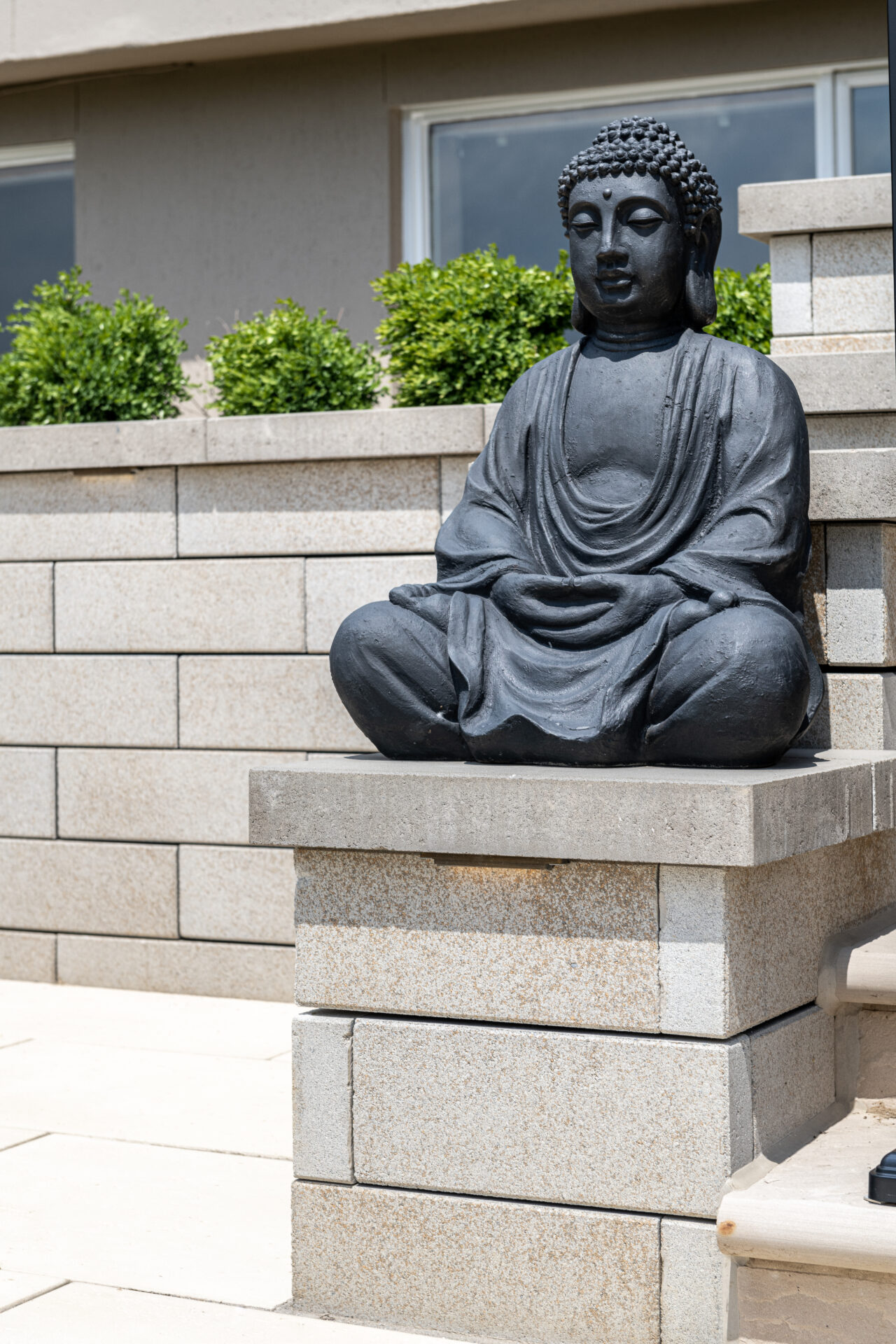 A Buddha statue sits peacefully on a modern, stone platform, surrounded by green shrubs and a simple wall backdrop.