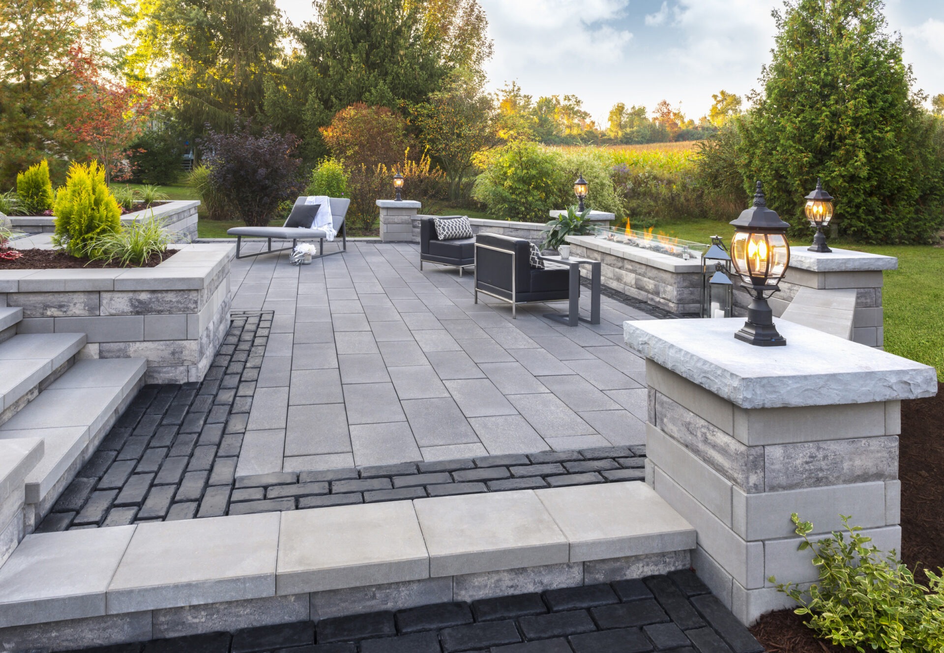 Modern patio with stone tiles, fire feature, and cushioned seating. Surrounded by green trees and lamp posts providing ambient lighting.