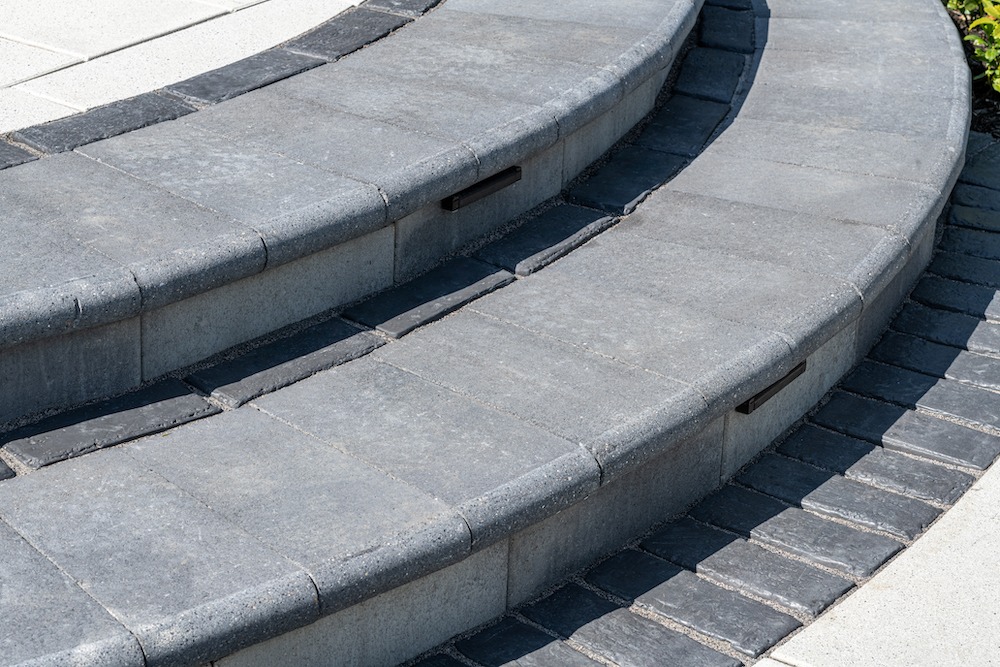 Curved gray stone steps with integrated lighting. The stairway is outdoors, featuring a clean and modern design with neatly arranged paving stones.