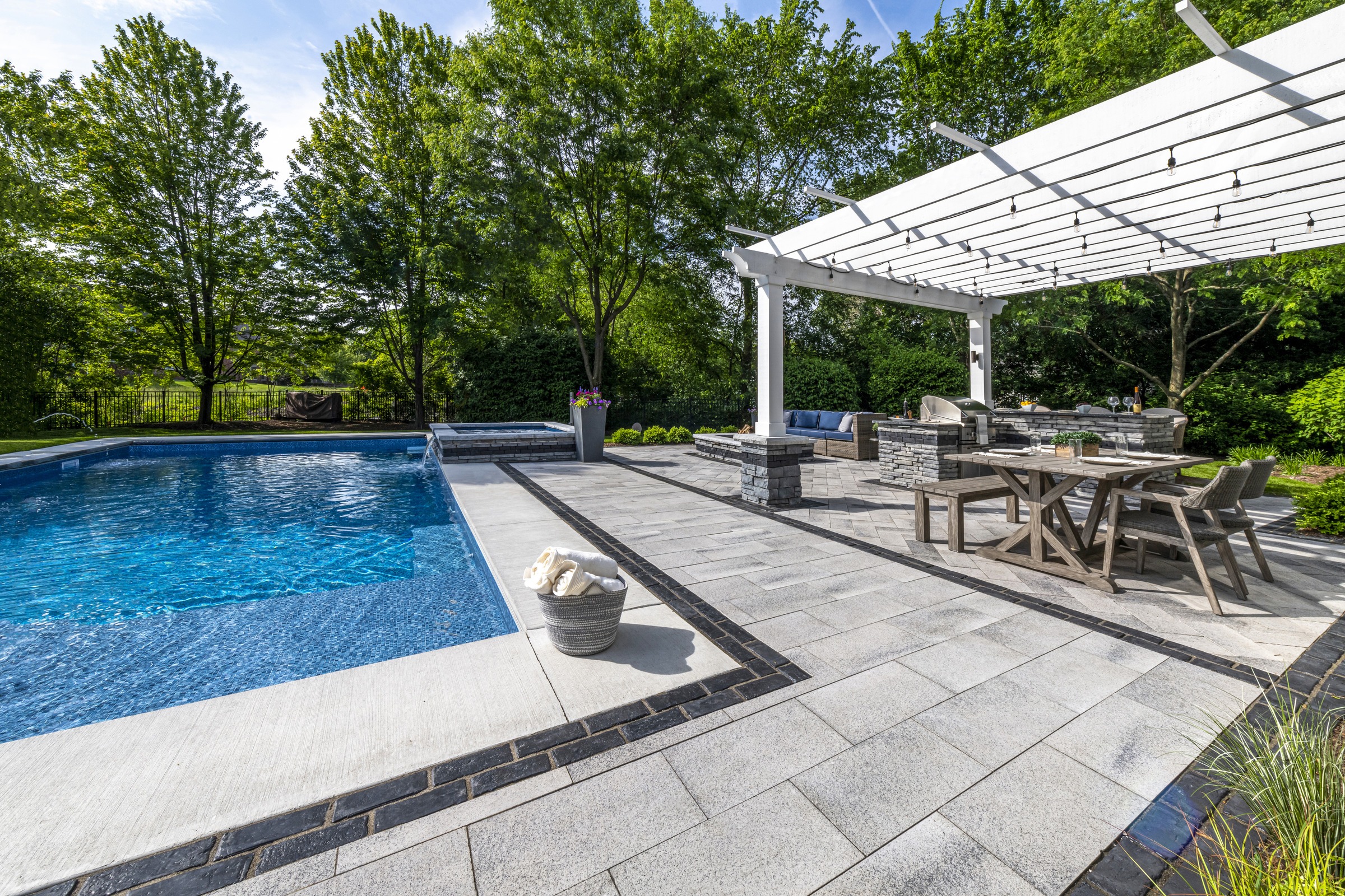 A modern patio with pergola, outdoor kitchen, and dining area beside a swimming pool, surrounded by lush green trees.