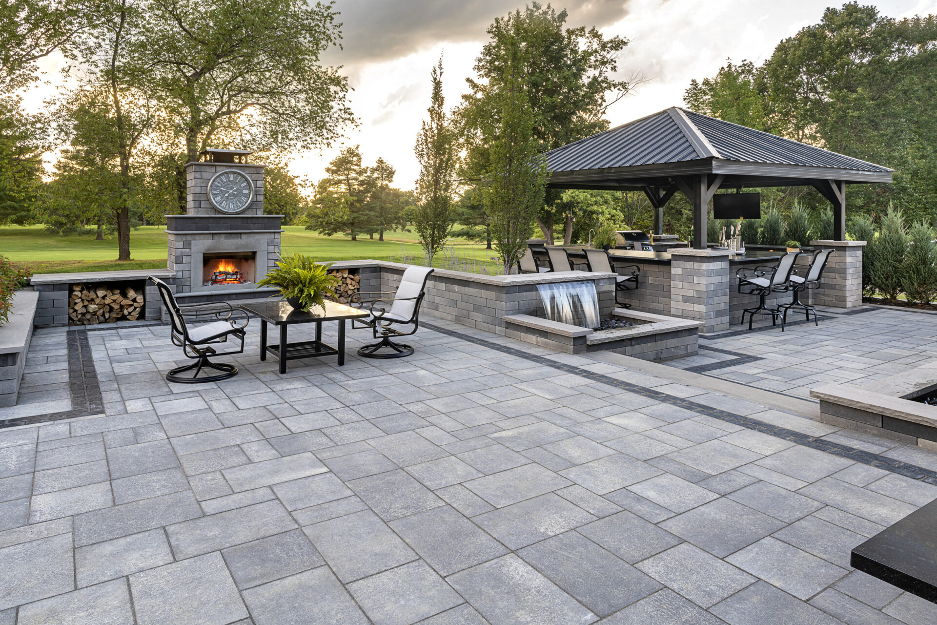 A modern outdoor patio features a fireplace, furniture, and a pergola-covered grilling area, surrounded by greenery and a serene park view.