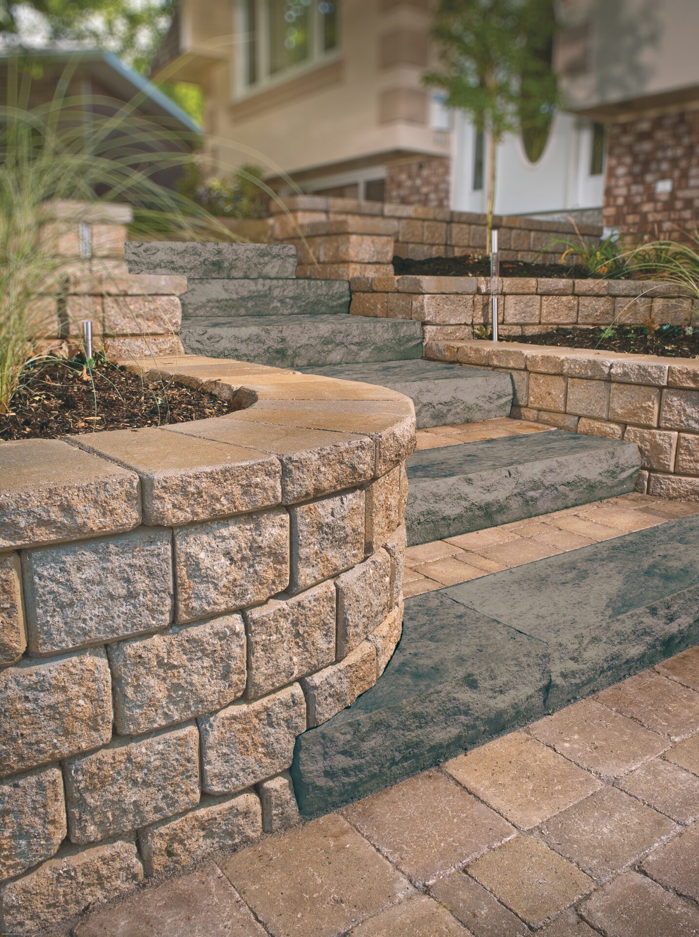 Stone steps and walls leading to a house entrance, surrounded by neatly arranged plants and greenery, creating a welcoming pathway.